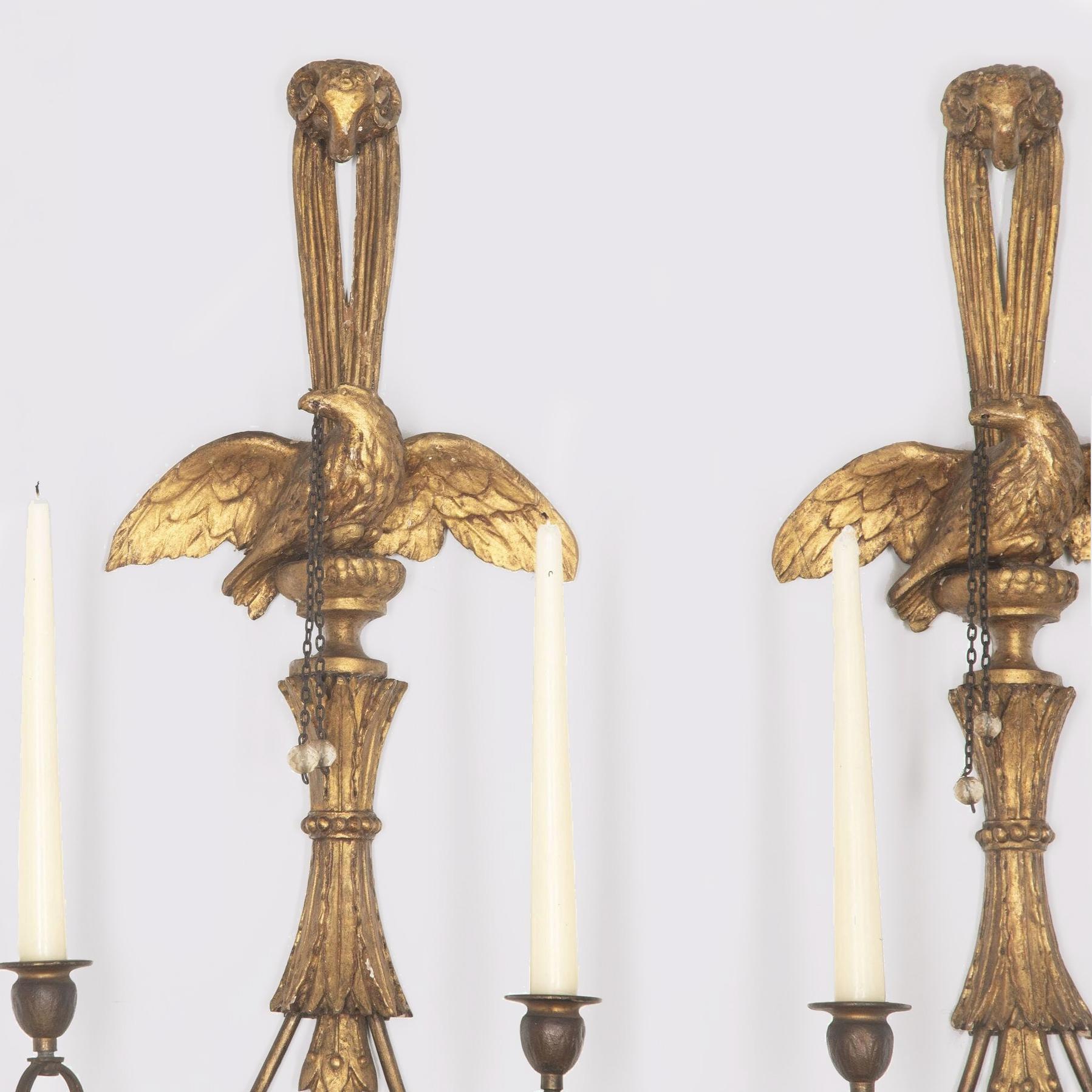 Finely carved pair of twin-branch English George III gilded wall lights. 

The carved ram's head above an eagle with swept wings holding a chain with a cut glass crystal bead. 

The gilt metal arms with leaf decoration.

