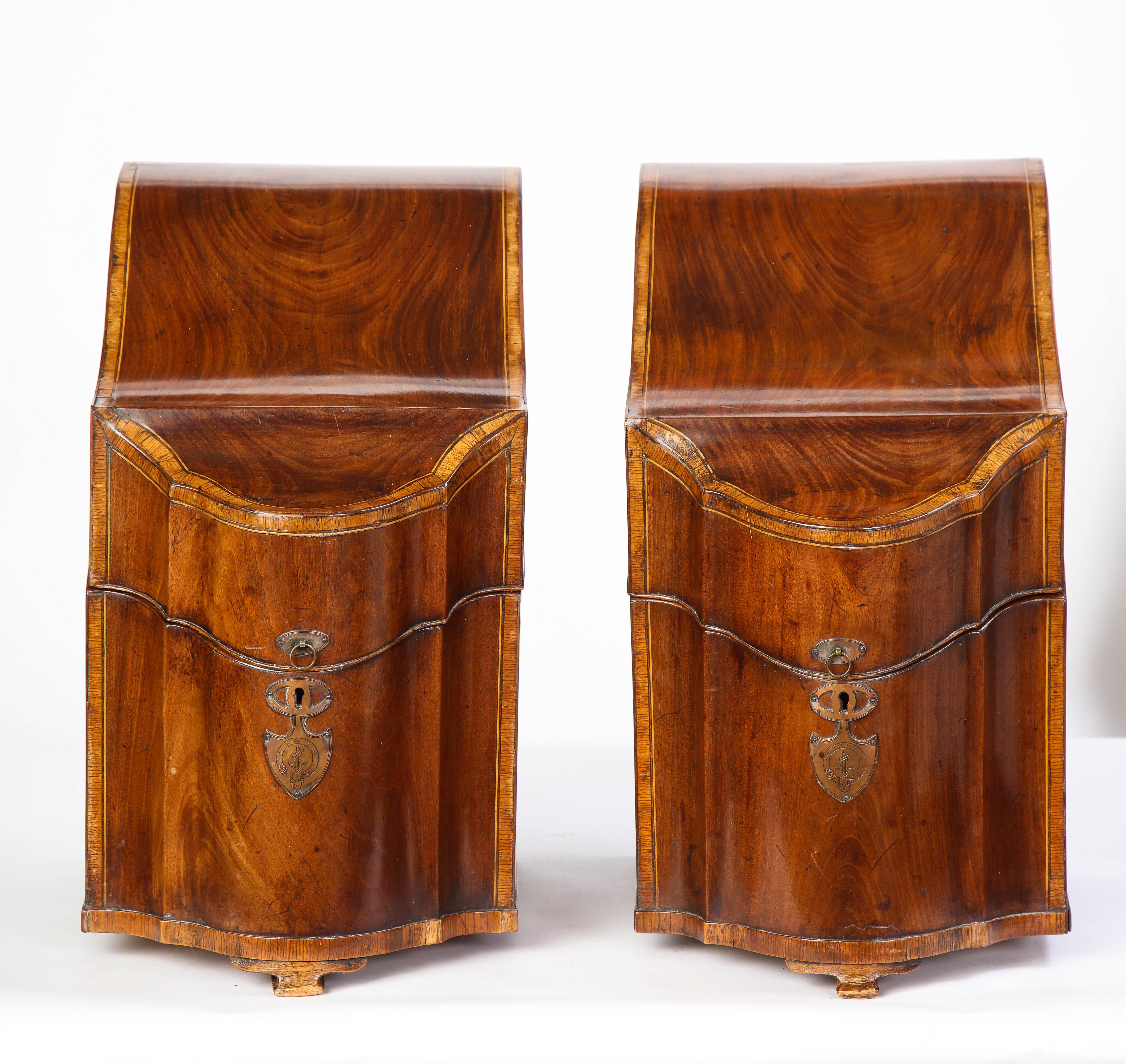 A matched pair of George III inlaid mahogany cutlery boxes from the late 18th century. Each box has a sloping serpentine-front hinged top enclosing a fitted wooden interior for flatware and cutlery. Both boxes boast a copper-tone metal escutcheon