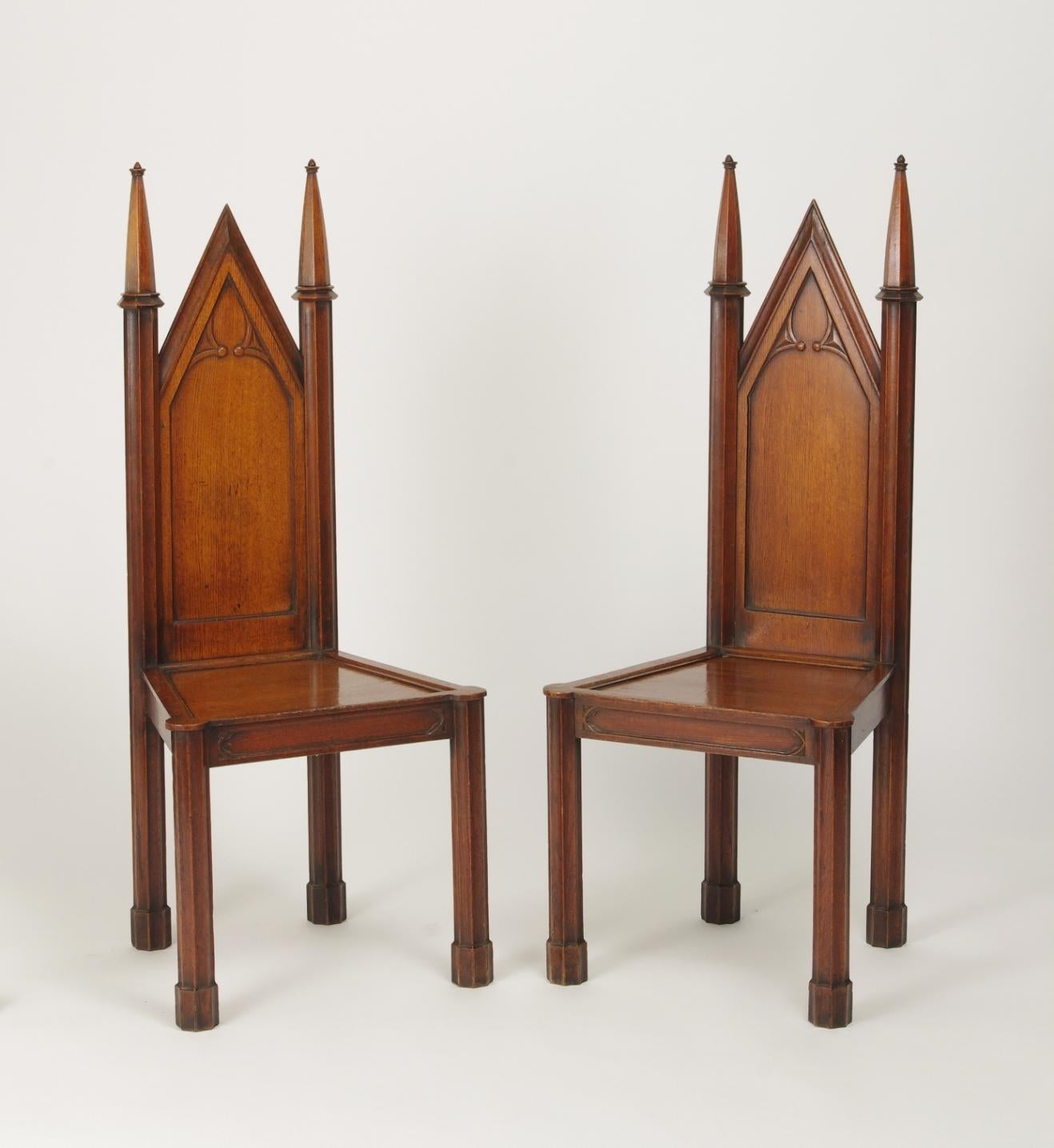 Pair of George III oak gothic hall chairs, each with a gabled back with gothic tracery between fluted spires, the plank seat with a molded edge on an apron with similar tracery and raised on fluted legs with plinth feet.

These stylish chairs,
