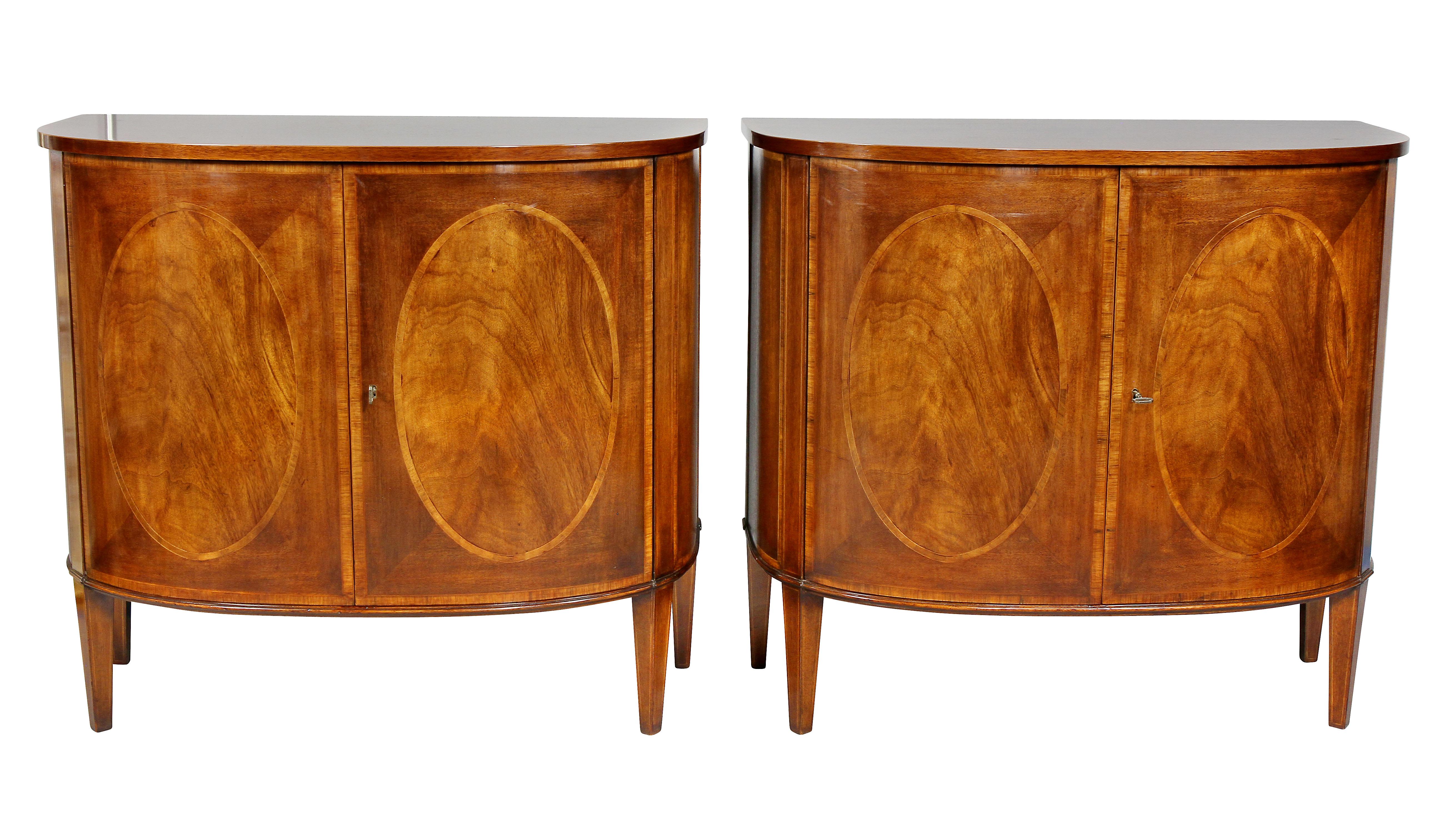 Each with a bowed crossbanded top over a pair of conforming doors with oval panels enclosing a shelf and raised on square tapered legs. Made by Smith and Watson, New York.