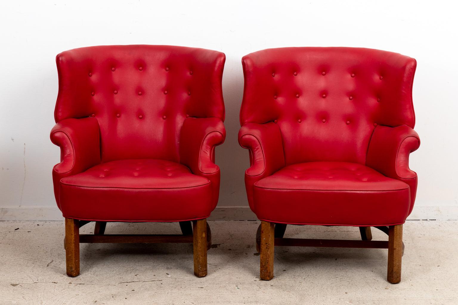 A pair of George III style wing chair upholstered in red vegan leather. The stretcher base follows the contour of the curved back. Two button covers missing on seat of one chair. Wear consistent with age.