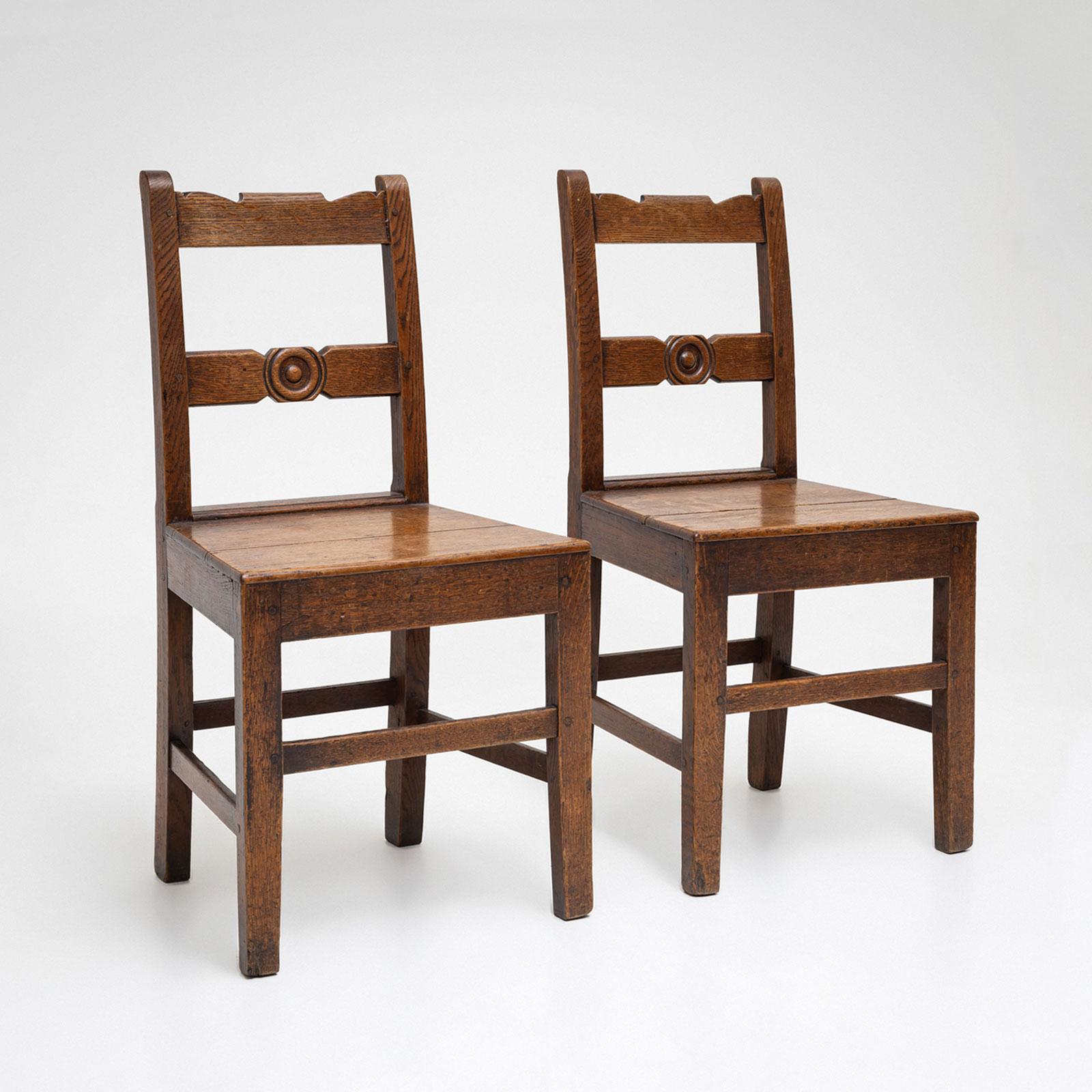 Paire de chaises provinciales George III en chêne massif, datant de la fin du XVIIIe siècle. Les chaises présentent des tabliers droits, des piétements robustes et des dossiers simples. L'éclisse centrale de chaque dossier est ornée d'un médaillon