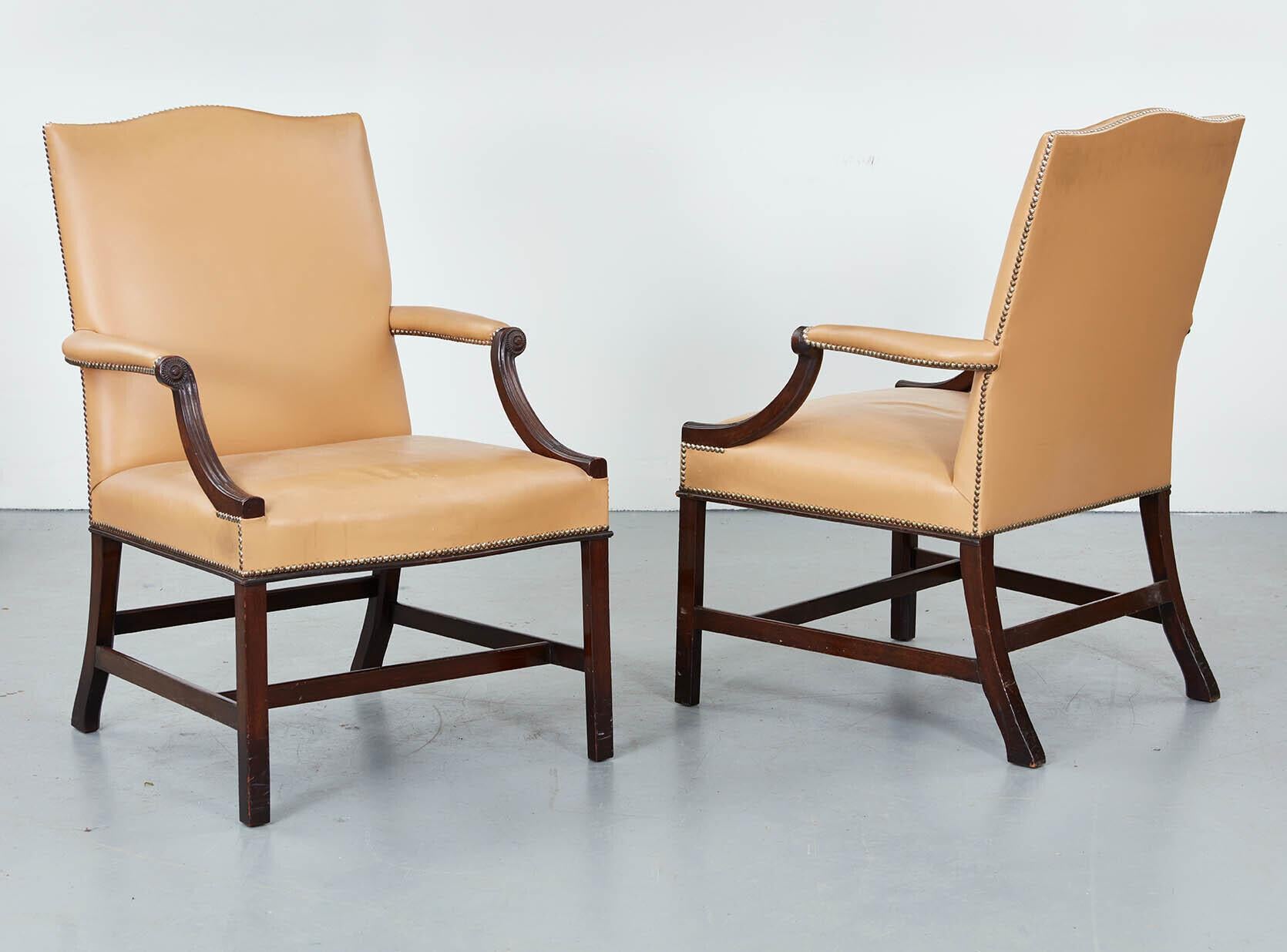 A pair of 1920’s Georgian style Gainsborough chairs in mahogany upholstered in beige leather with close-nailed tacks, having molded down swept arm supports and standing on carved and molded legs joined by stretchers, having good color and great