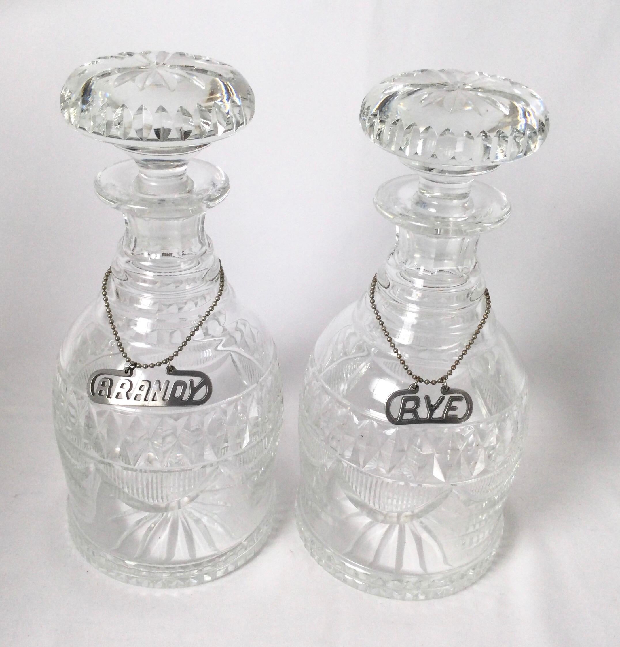 A pair of handcut glass liquor decanters with original stoppers. The bottles with ring necks, diamond pattern cut band and draped decoration. The bottoms with a starburst pattern in the center. Mid-later 19th century with a later pewter label on