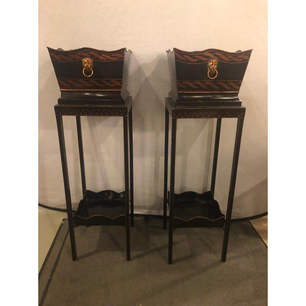 A beautiful pair of Georgian style tole hand painted jardinières or planters featuring black, brown and rose gold colors in a wreath decorated pot. The pair of the planter offer an extra shelf and hand carved lion handles adding sophistication.
