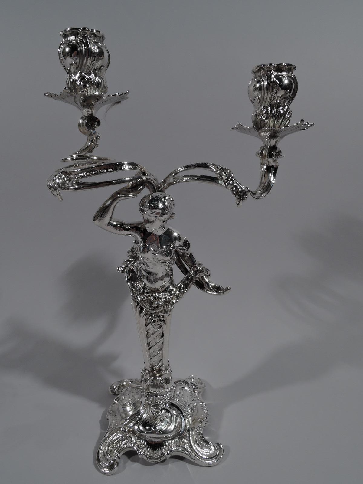 Pair of turn-of-the-century 800 silver 2-light candelabra. Made by Made by M. H. Wilkens & Söhne in Germany. Each: Tapering pillar on raised shaped foot with spread C-scroll supports. Garland-entwined figure mounted to pillar top. On one, a bosomy,