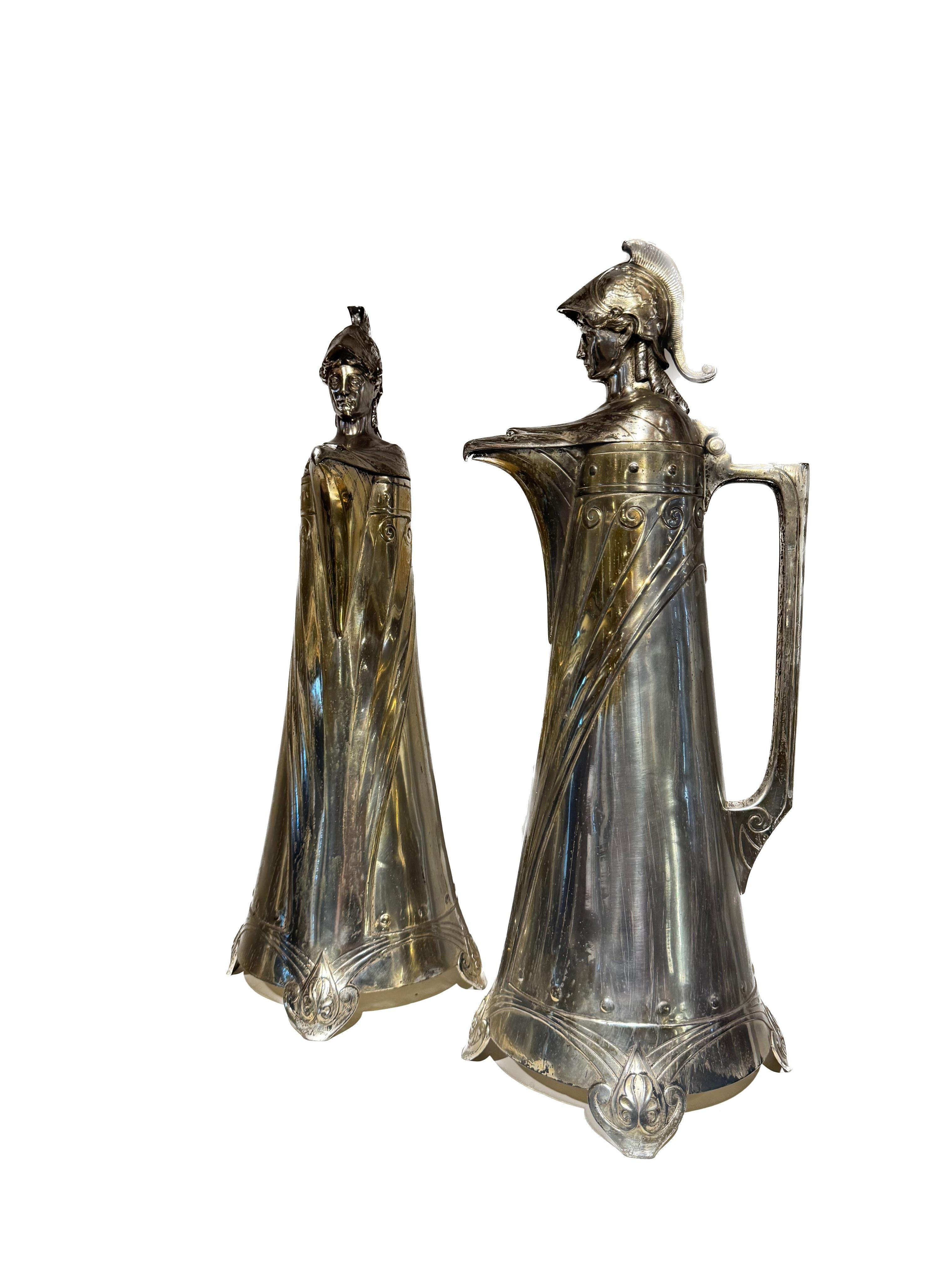 Un'impressionante coppia di brocche in metallo tedesche WMF (Württembergische Metallwarenfabrik) in stile Art Déco, ognuna delle quali presenta intricati disegni di un soldato in rilievo e dettagliati motivi a testa d'aquila. Questi pezzi decorativi