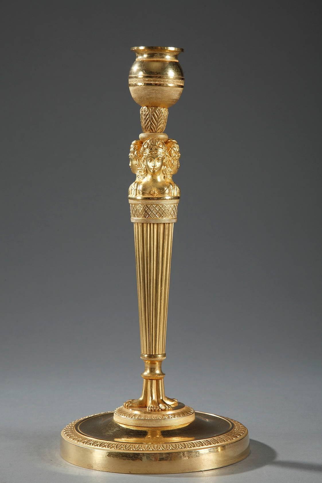 Paar vergoldete und skulptierte Bronzeleuchter aus der Empirezeit. Der Sockel ist sehr aufwändig mit Perlen und Blattwerk modelliert. Unter dem Sockel sind drei nach außen gerichtete Frauenbüsten zu sehen, die den oberen Teil des kannelierten,