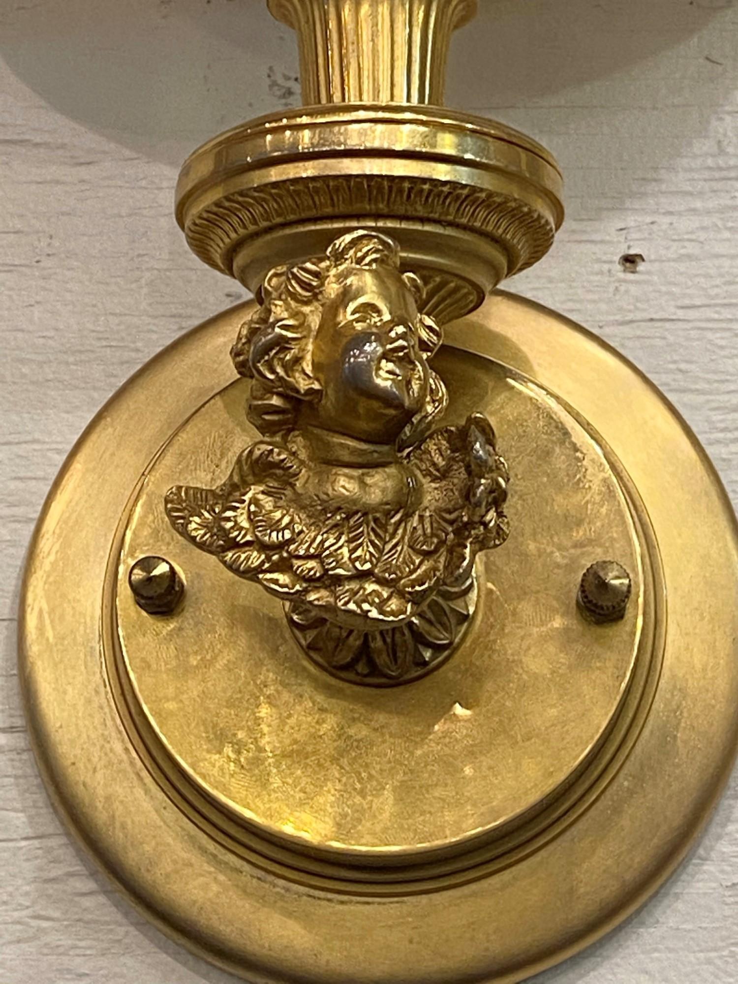 This pair of wall sconces are gold gilded over brass, and are designed with a cherub supporting a single arm. The original white shade is included. They can be seen at our 2420 Broadway location on the upper west side in Manhattan.