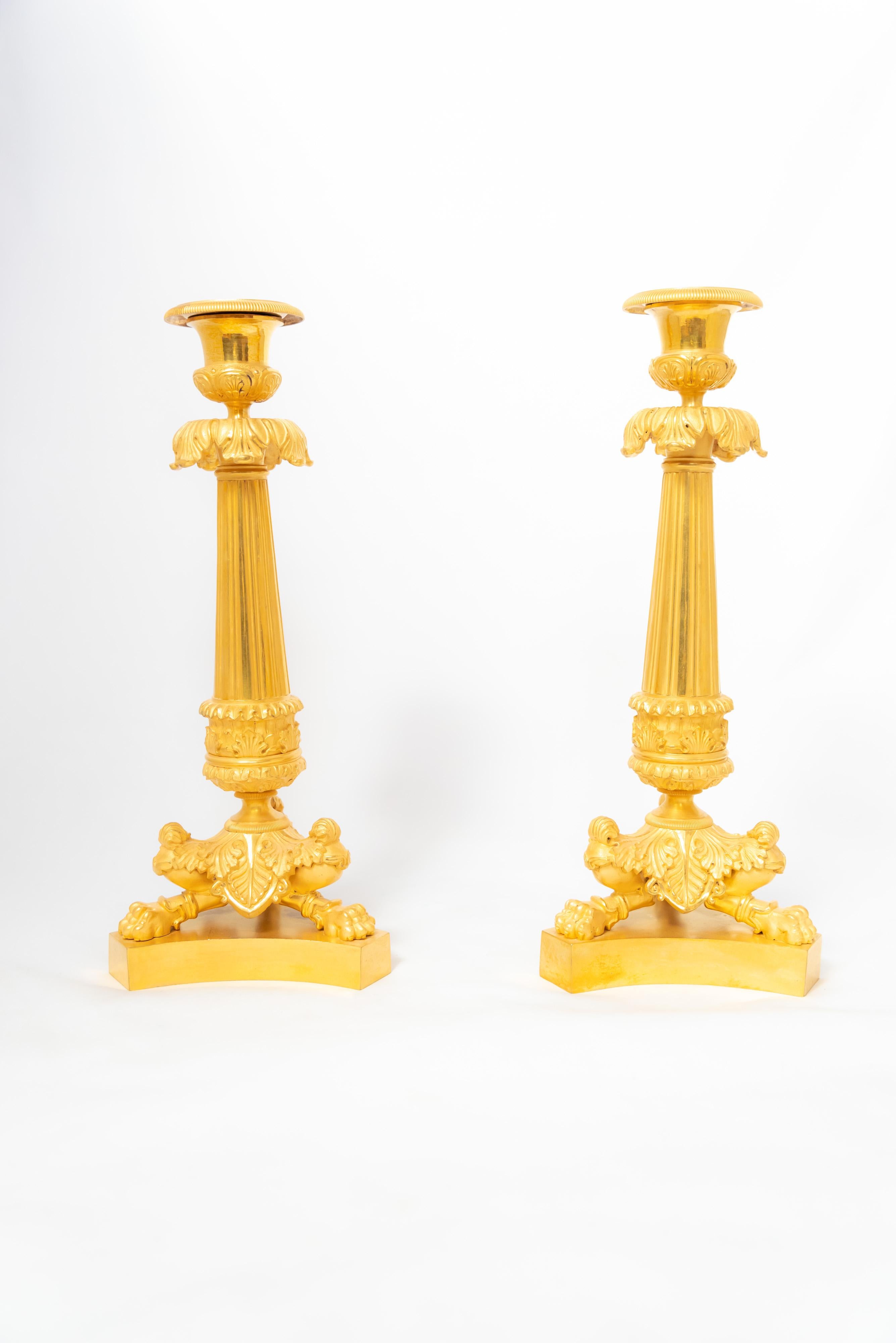 A pair of candlesticks in chiselled fire-gilt bronze from the French Empire or early Restauration Era. The pair, exemplifying the neoclassical iconography so popular in the Napoleonic Age, features a lion’s-paw-tripod base on a plinth complemented