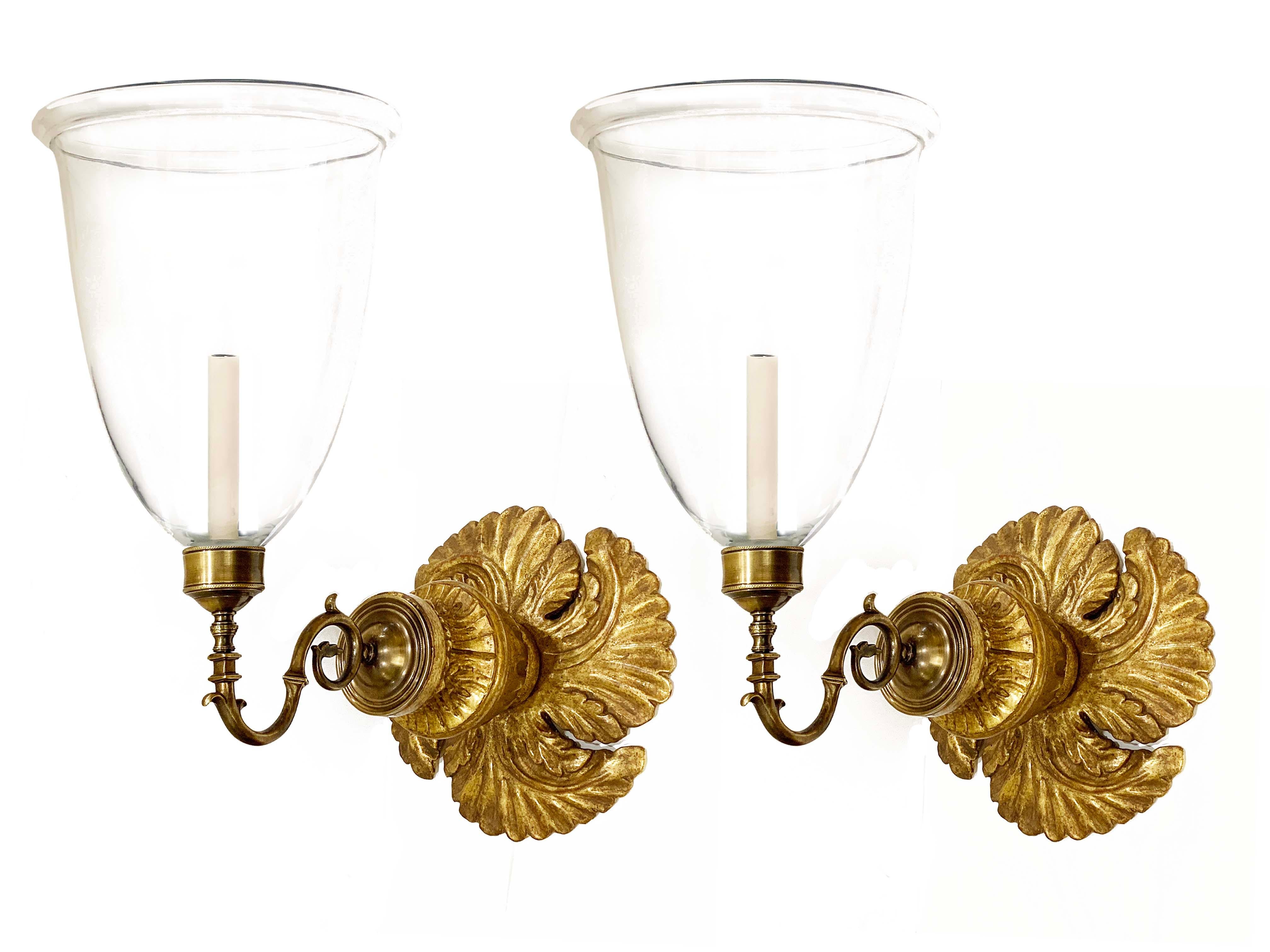 A pair of hand-carved gilt Georgian-style backplates with patinated brass sconce fittings and large hand-blown clear glass hurricane shades. Electrified with one candelabra base socket per sconce.

Our custom hurricane sconces are available with
