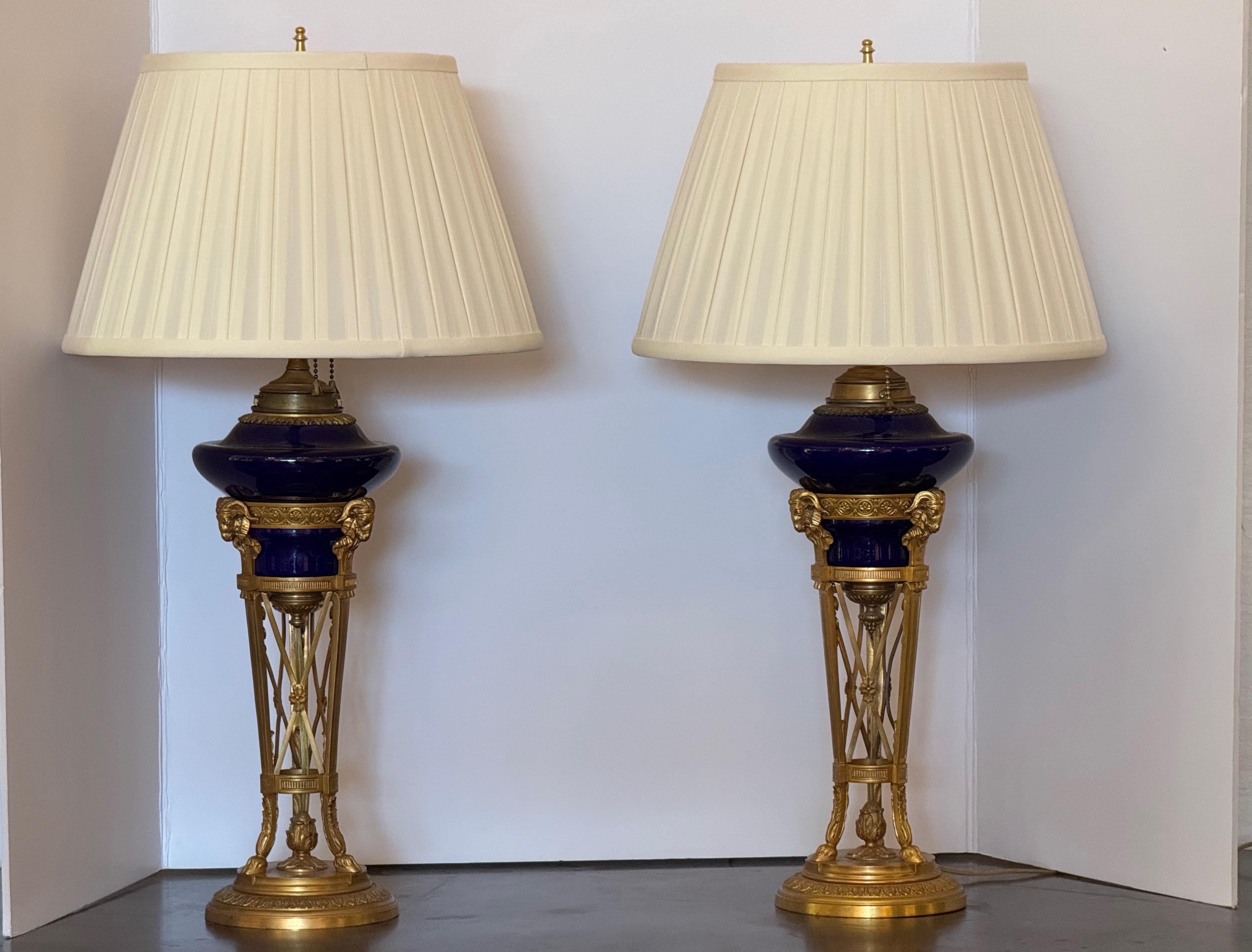 Pareja de lámparas de sobremesa de estilo neoclásico con cuerpos de cristal azul cobalto engarzados en elaboradas monturas de bronce dorado. Cada lámpara se alza sobre una base redonda con detalles de acanto y guirnalda, que se eleva hasta una