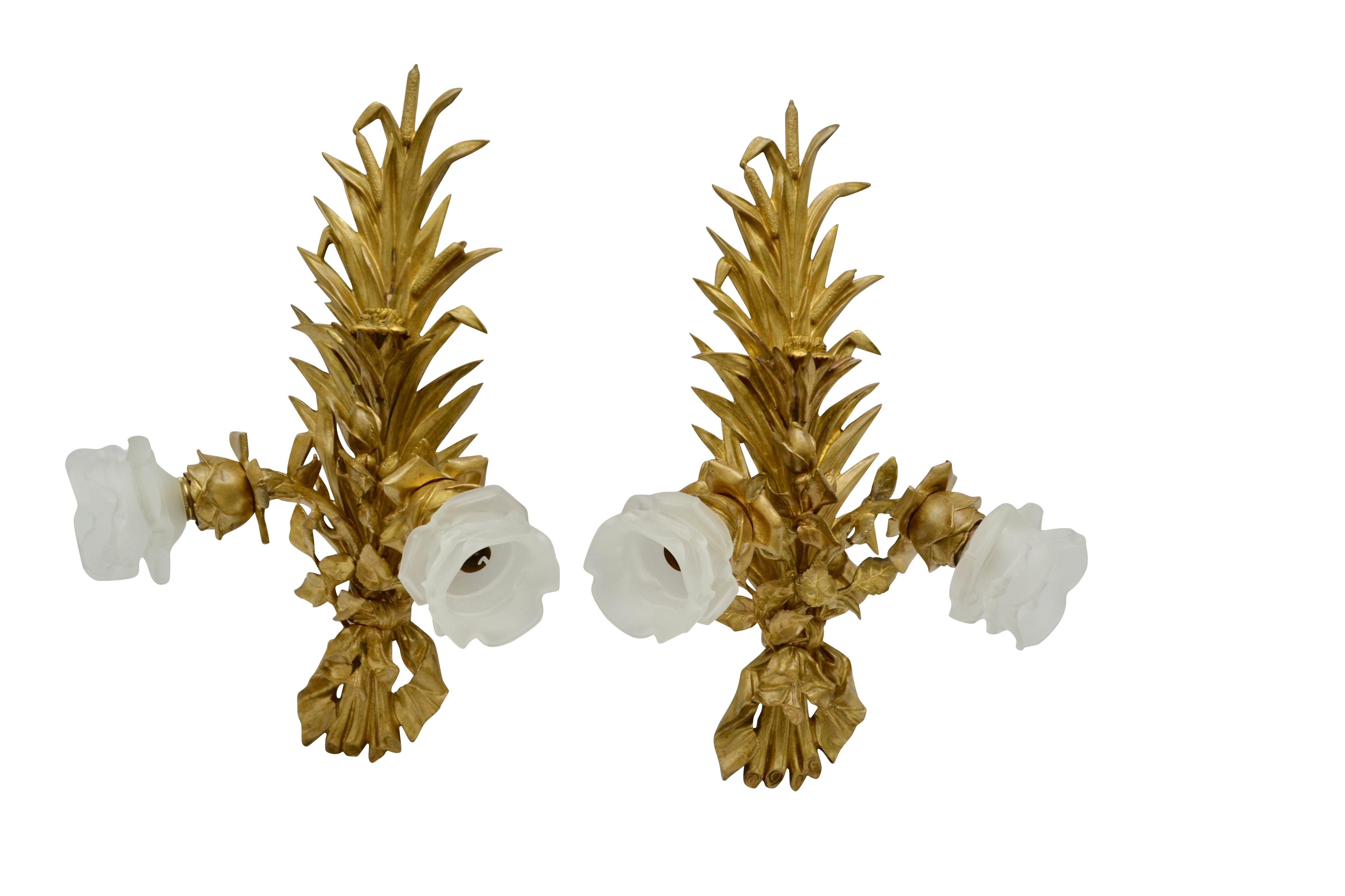 A pair of very well cast wall sconces in the shapes of bullrushes. The bronzes show signs of earlier gilding and are now a dull gold colour. Each sconce has a pair of electrified decorative floral nozzles holding a frosted glass flower shade. There