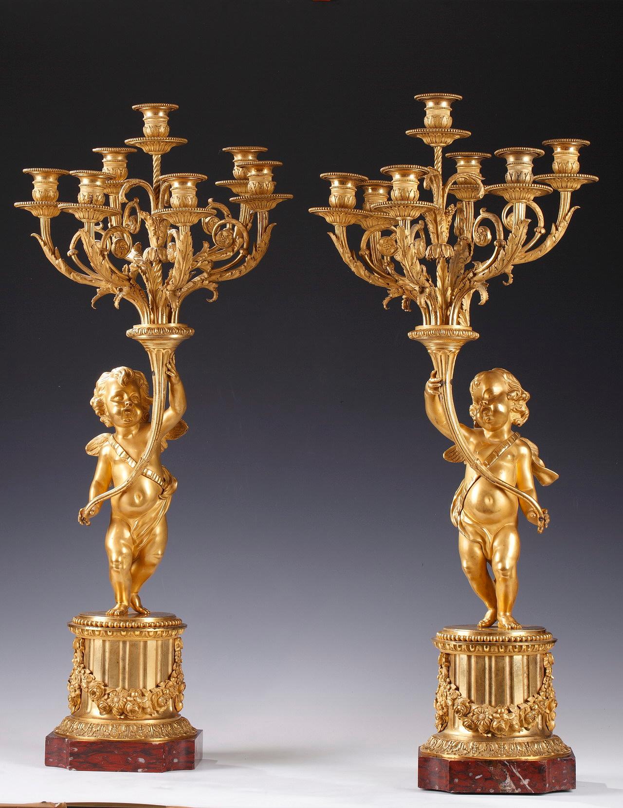Large pair of candelabras after a model by Clodion, representing two winged putti in gilded bronze, each holding a cornupia containing seven light-arms adorned with foliage. They rest on a square base with curved edges in Griotte marble, surmounted