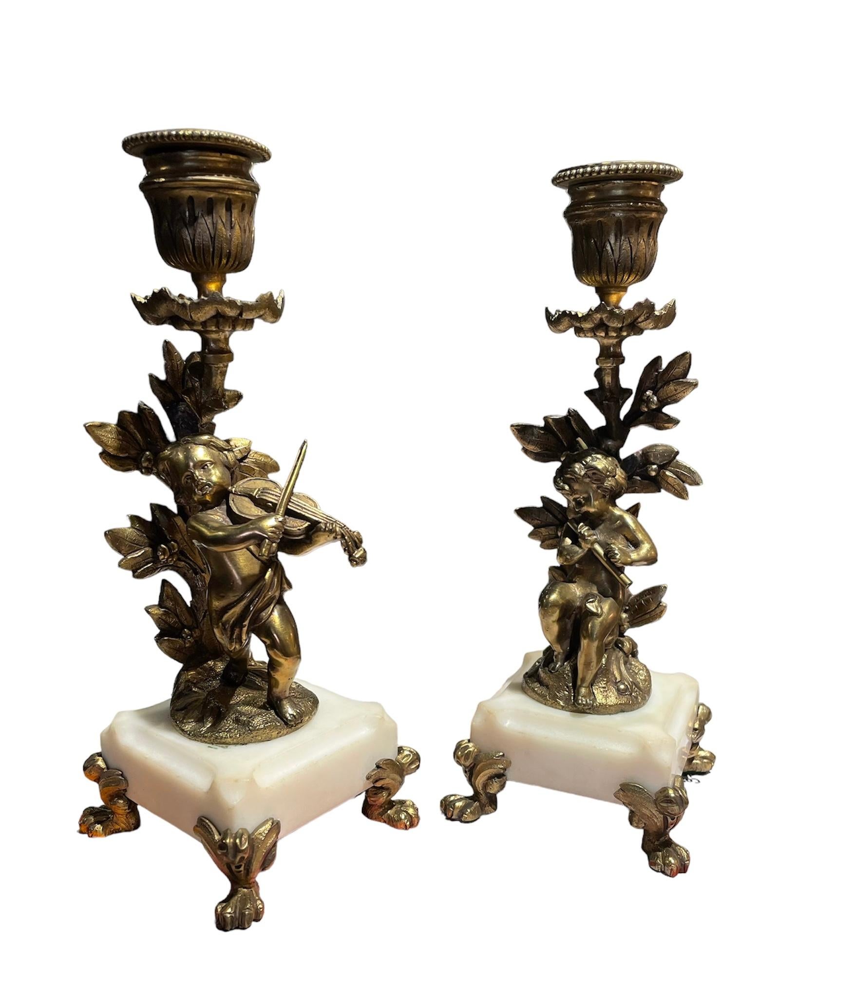 This is a pair of gilt bronze candle holders. Each one of them depicts a cherub figure who is playing an instrument in front of a bay leaves tree. One is playing the violin and the other one the flute. From one of the bay leaves branches come out
