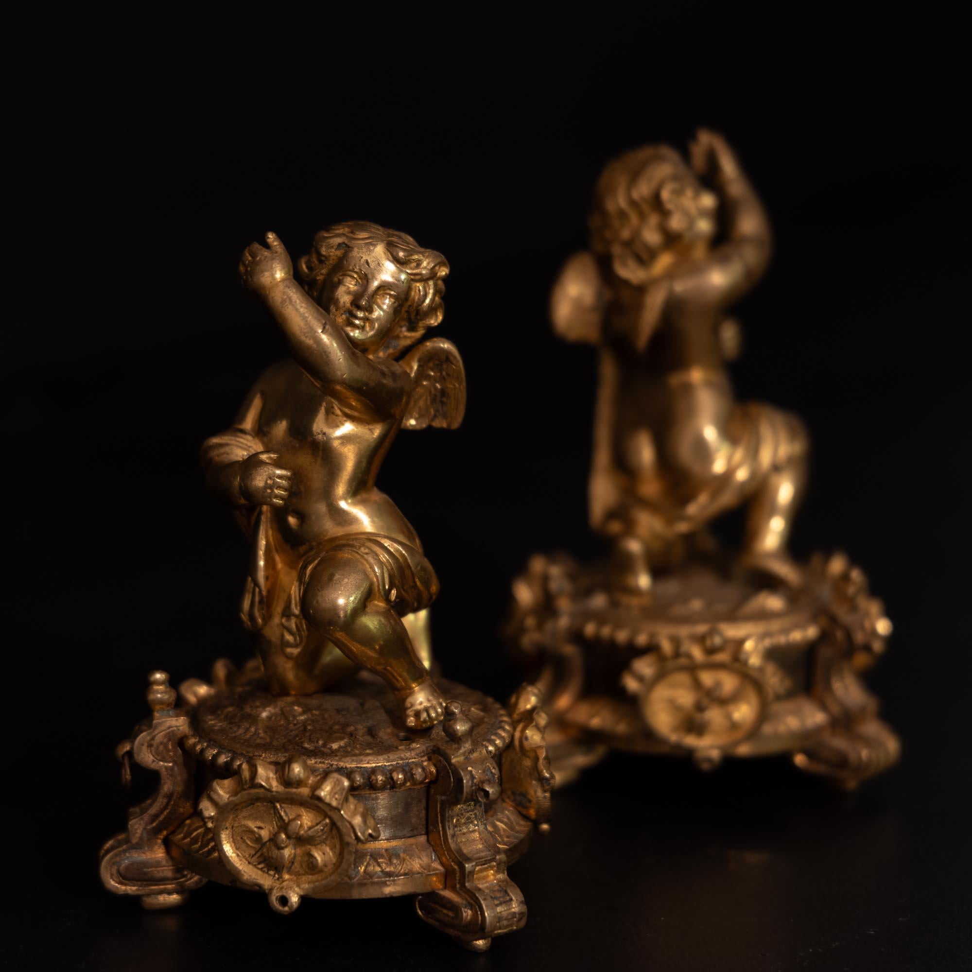 Pair of gilt bronzes in the form of winged putti with raised arms and opposite design. The putti stand on round bases with volute finials, pearl staff and medallions. The putti originally carried something in their arms, a small hole in the base of