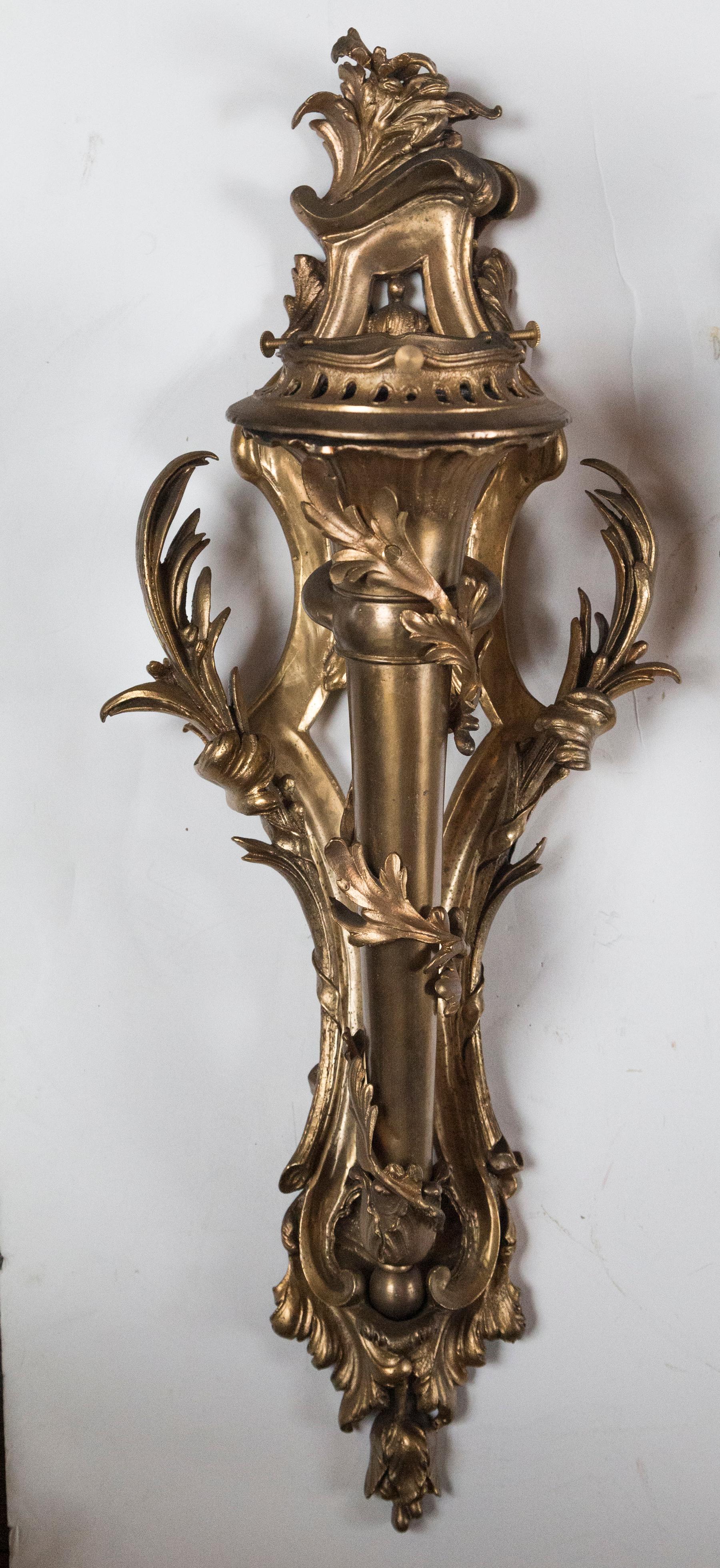 The gilding in antique gold color. Each in the form of a torch., encircled with an wreath of acanthus leaves. Leaf adornments to the top and bottom, with splayed out leafy arms on either side. The central mounting is part of the sconce. unwired.