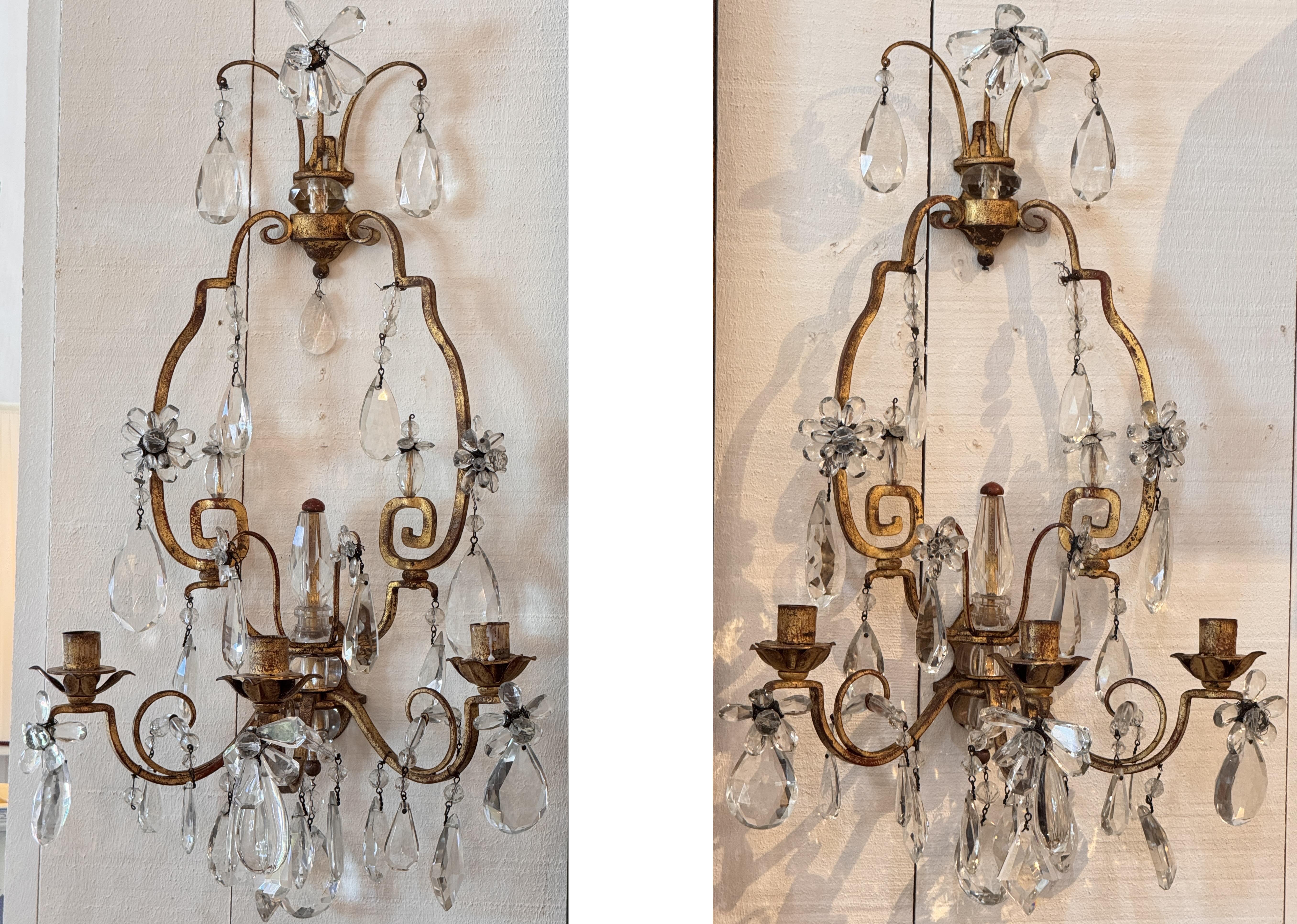 Ein Paar vergoldete Metallleuchten mit geschliffenen Kristallprismen und Blumenrosetten. Die geschwungenen Arme tragen jeweils drei Kerzenlichter, während die vergoldeten Rahmen mit Tropfen und facettierten Kristallelementen akzentuiert sind. Zu den