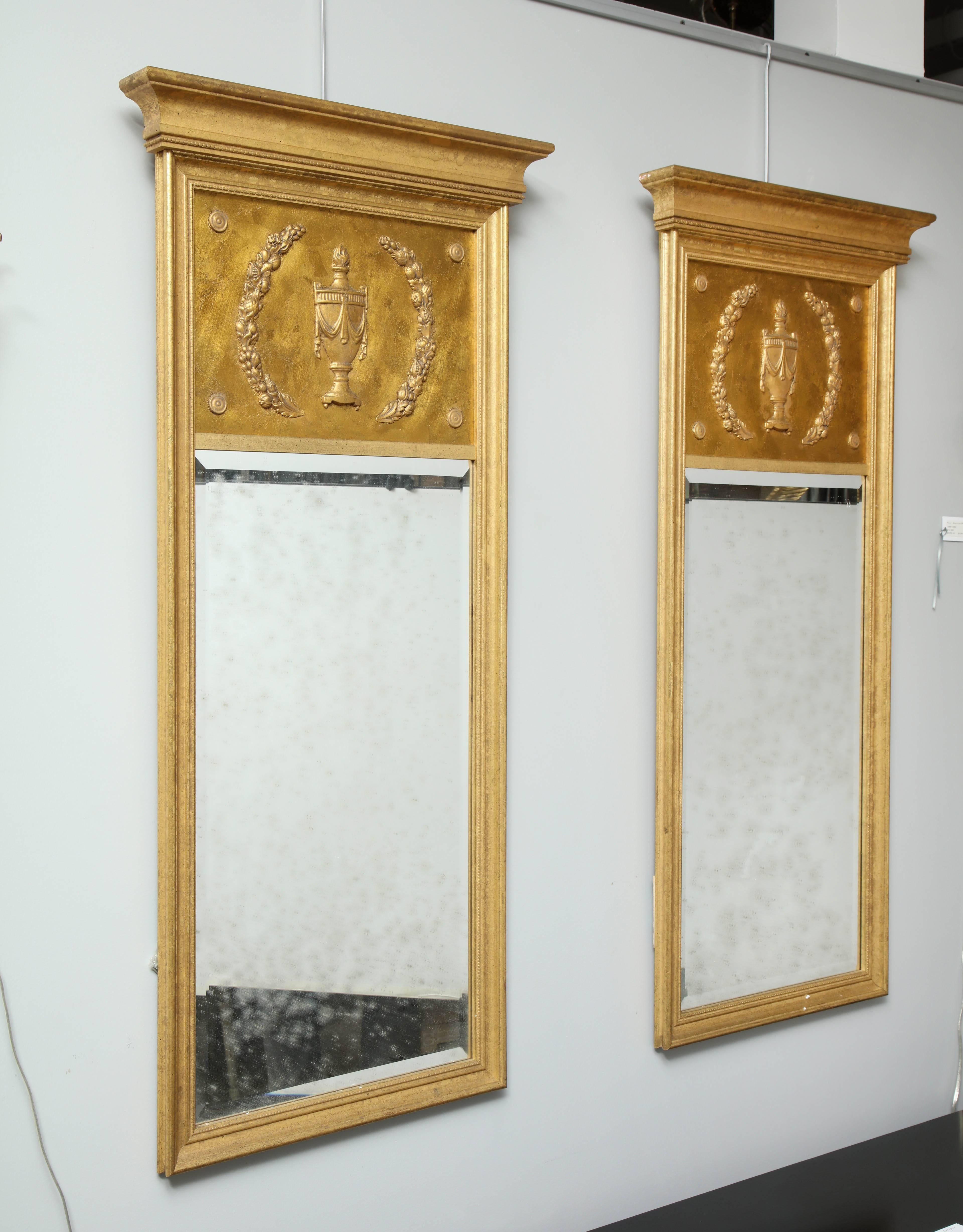 Una coppia di specchi in legno dorato, sobri ed eleganti. La cornice modellata circonda il vetro smussato anticato e un motivo neoclassico intagliato sulla parte superiore.