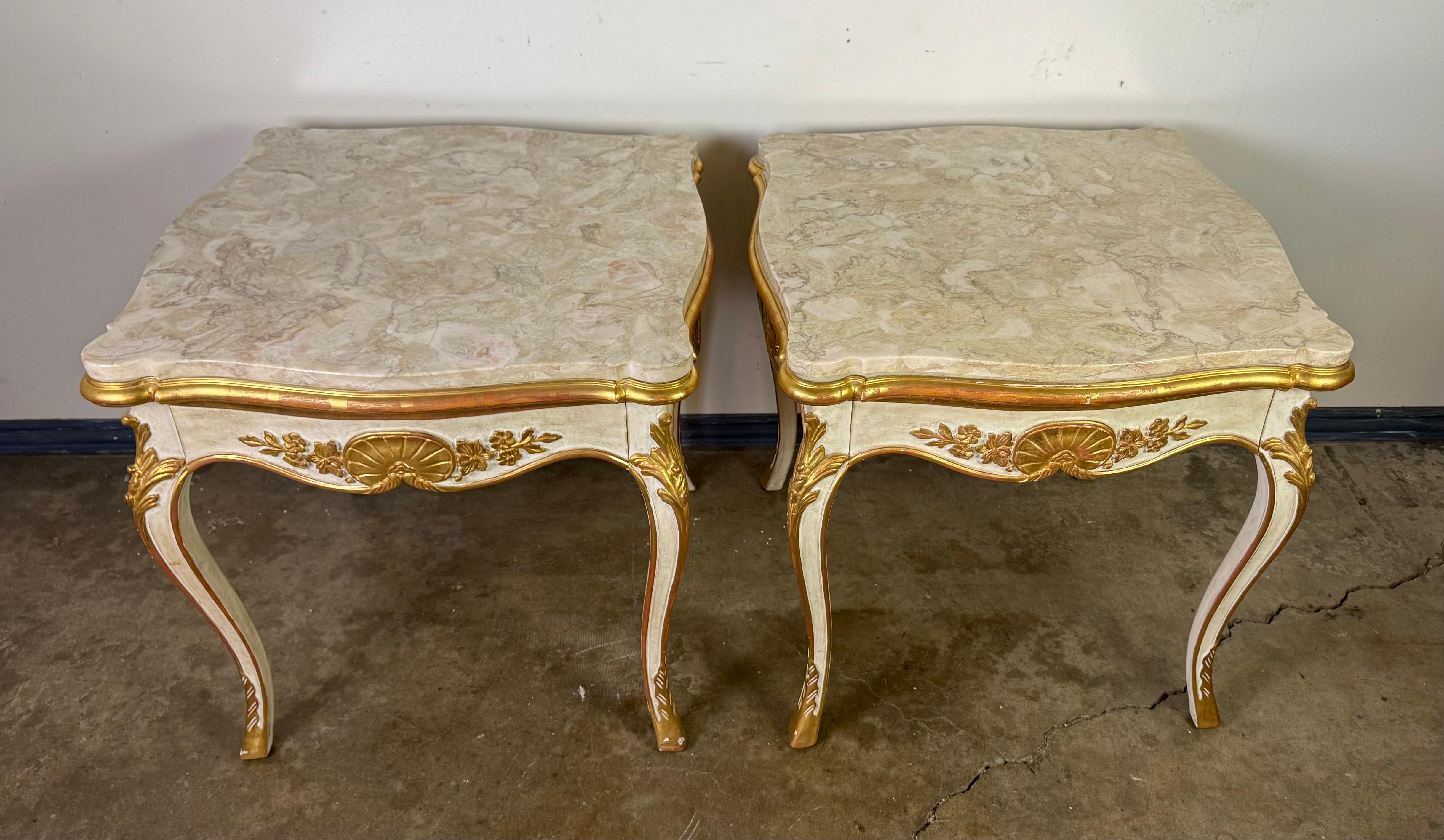 A striking pair of Rococo Revival��–style side tables, each with a single hidden drawer. The tables feature shaped faux-marble tops in soft cream and beige veining, resting over serpentine aprons with carved and gilt shell and floral motifs at the
