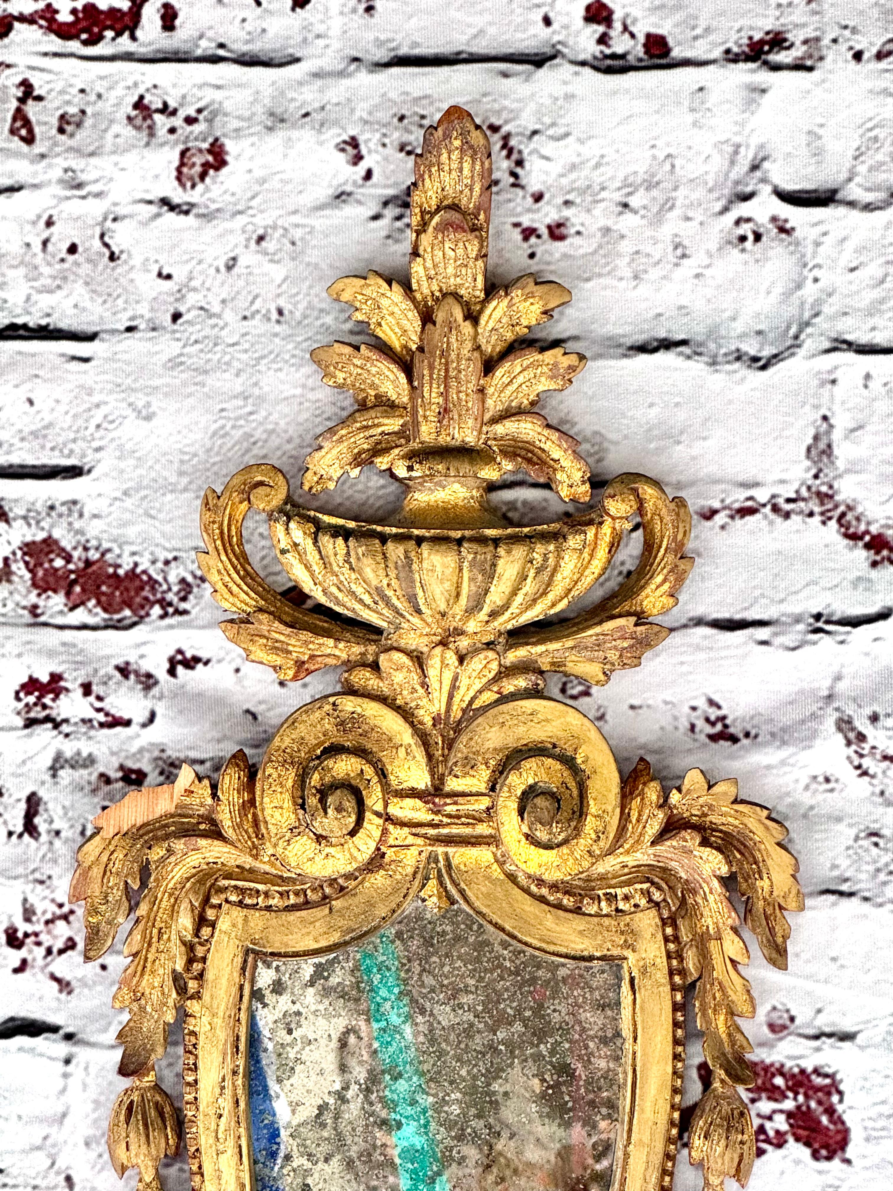 Superbe paire d'appliques murales italiennes en bois doré, chacune avec un miroir en forme de bouclier et deux bras avec des chapeaux de bougie singuliers. Les deux bras de lumière se reflètent dans les miroirs pour un aspect opulent. Chaque