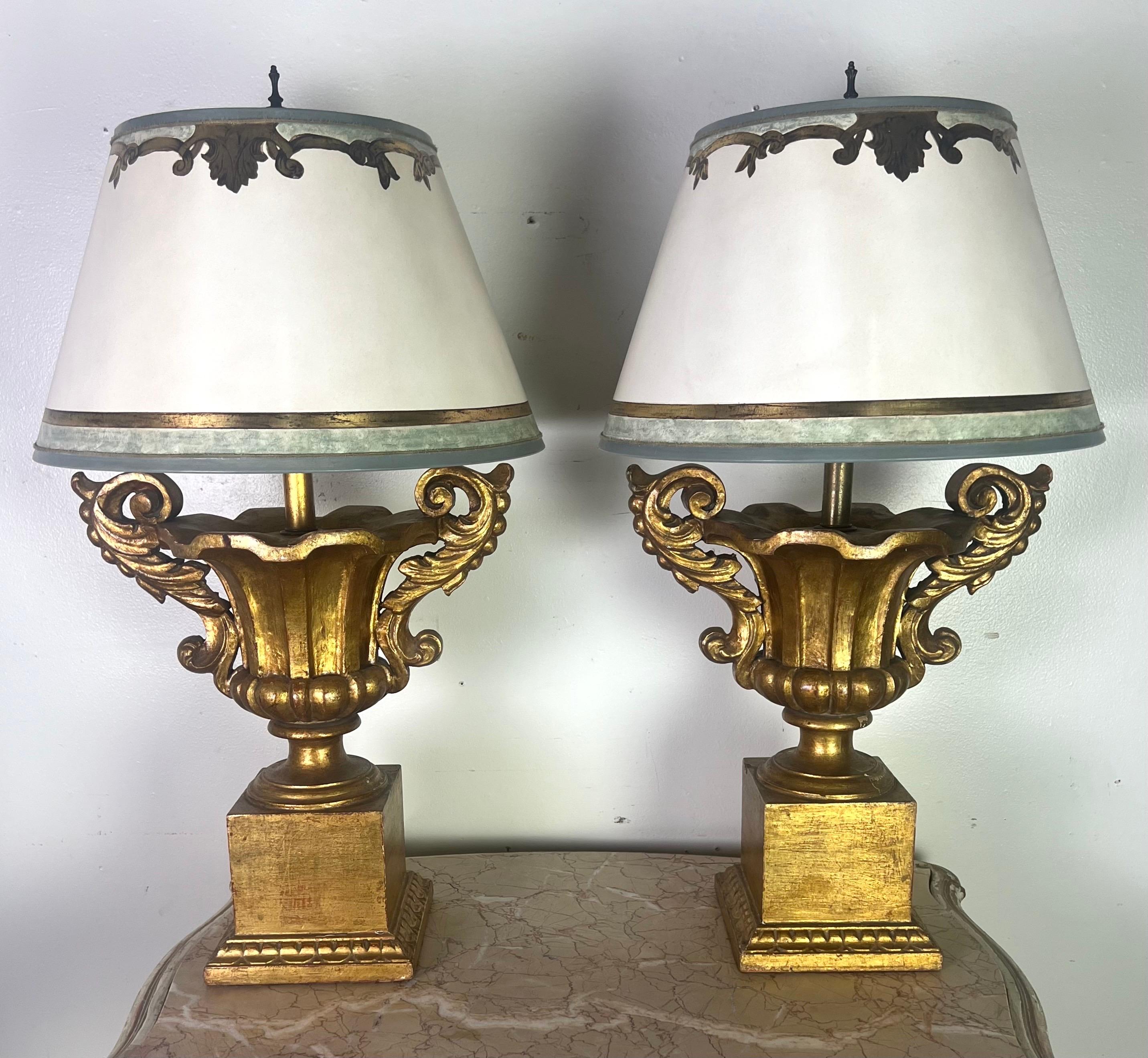 Coppia di lampade a urna in legno dorato italiano in stile neoclassico con paralumi in pergamena dipinti a mano. Le basi delle lampade sono in stile urna classica, con elaborate volute e dettagli decorati.  Sono rifiniti con una ricca foglia d'oro