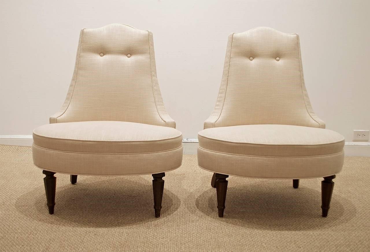 A pair of glamorous Hollywood Regency style tufted lounge chairs. The arm less chairs features a curved back, with an oval seat, standing on reeded front legs. The chair is upholstered in a cream silk fabric, with button tufting to back and seat.