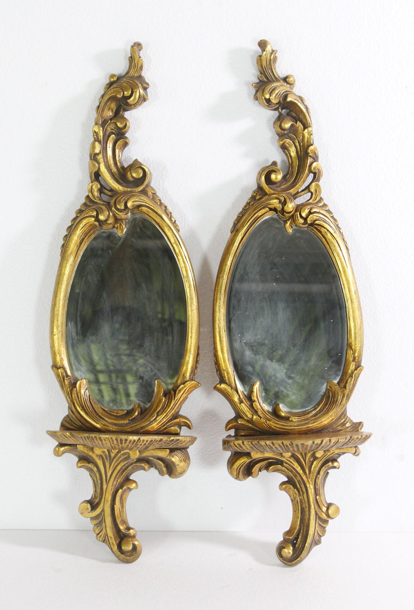 Paire de miroirs muraux en bois doré avec étagère et détails de feuillage. Les miroirs sont de forme ovale et le cadre en bois est orné de détails floraux sur l'ensemble du plateau et du dessus. Le prix est fixé par paire. Veuillez noter que cet
