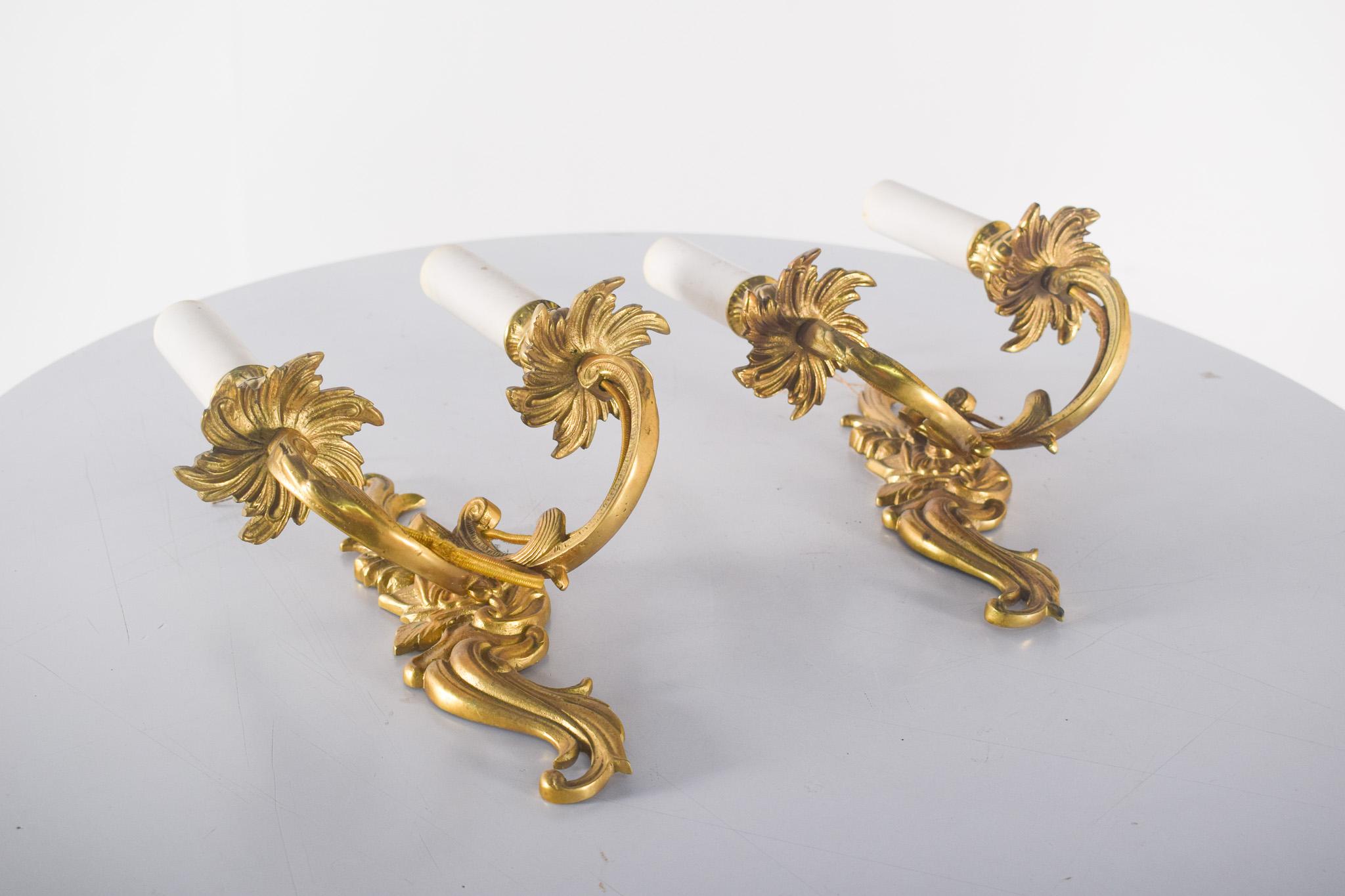 Pareja de apliques dorados. Decorado con volutas y hojas detalladas.  Cada aplique tiene dos brazos sinuosos que sostienen los portalámparas, simulando velas, que recuerdan la iluminación tradicional del siglo XVIII.
El acabado dorado resalta los