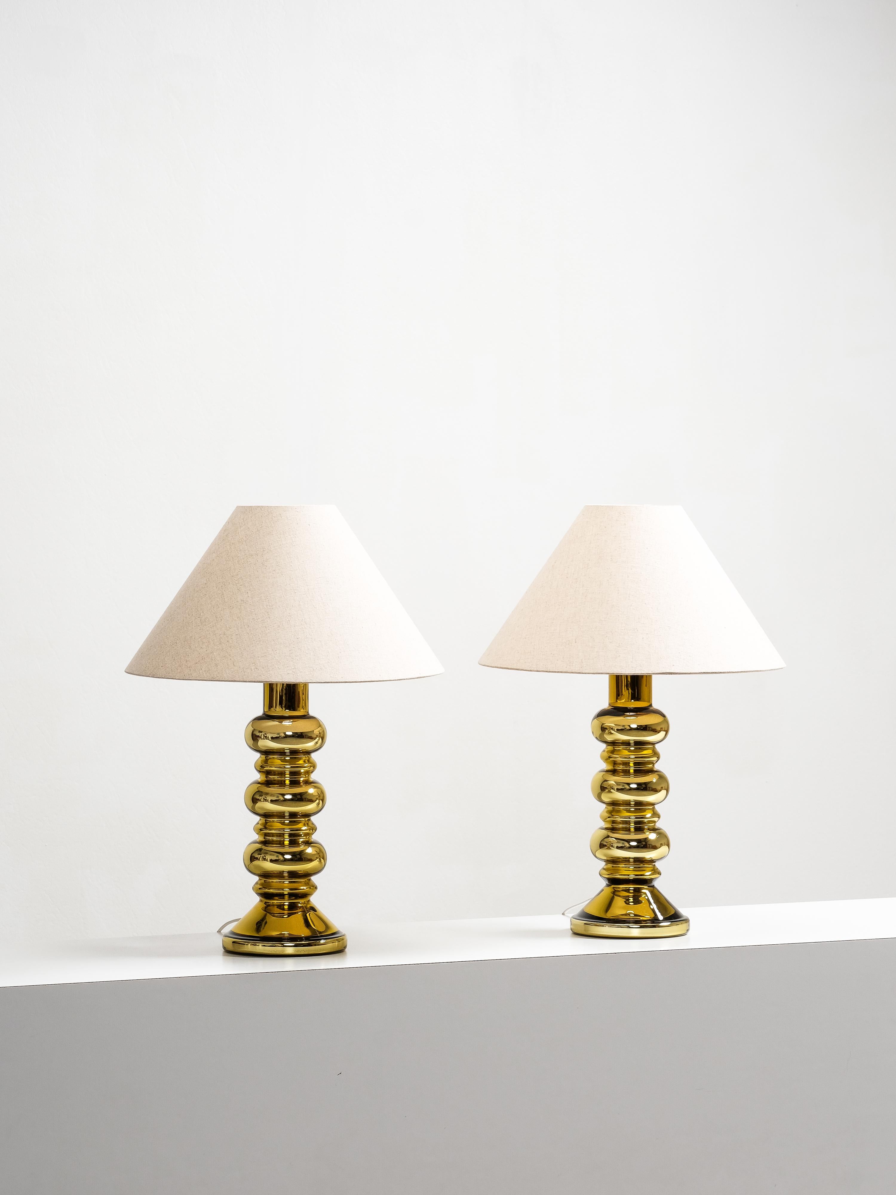 A pair of sculptural table lamps by Flygsfors, Sweden, from the 1960’s. The lamp bases are crafted in golden mercury glass with a bold stacked form, their mirrored surfaces reflecting and amplifying light. Each lamp is paired with a simple conical