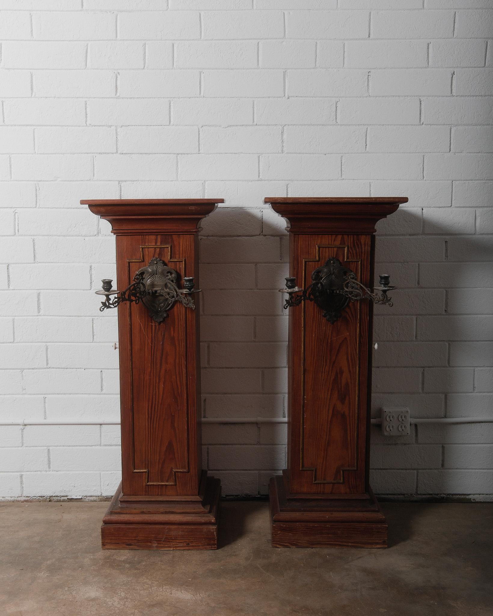 This stunning pair of Gothic Revival church pedestal stands features richly grained pine construction with beautifully molded details and inset paneling, enhanced by their original wrought iron candle sconces. These tall, stately pieces were likely