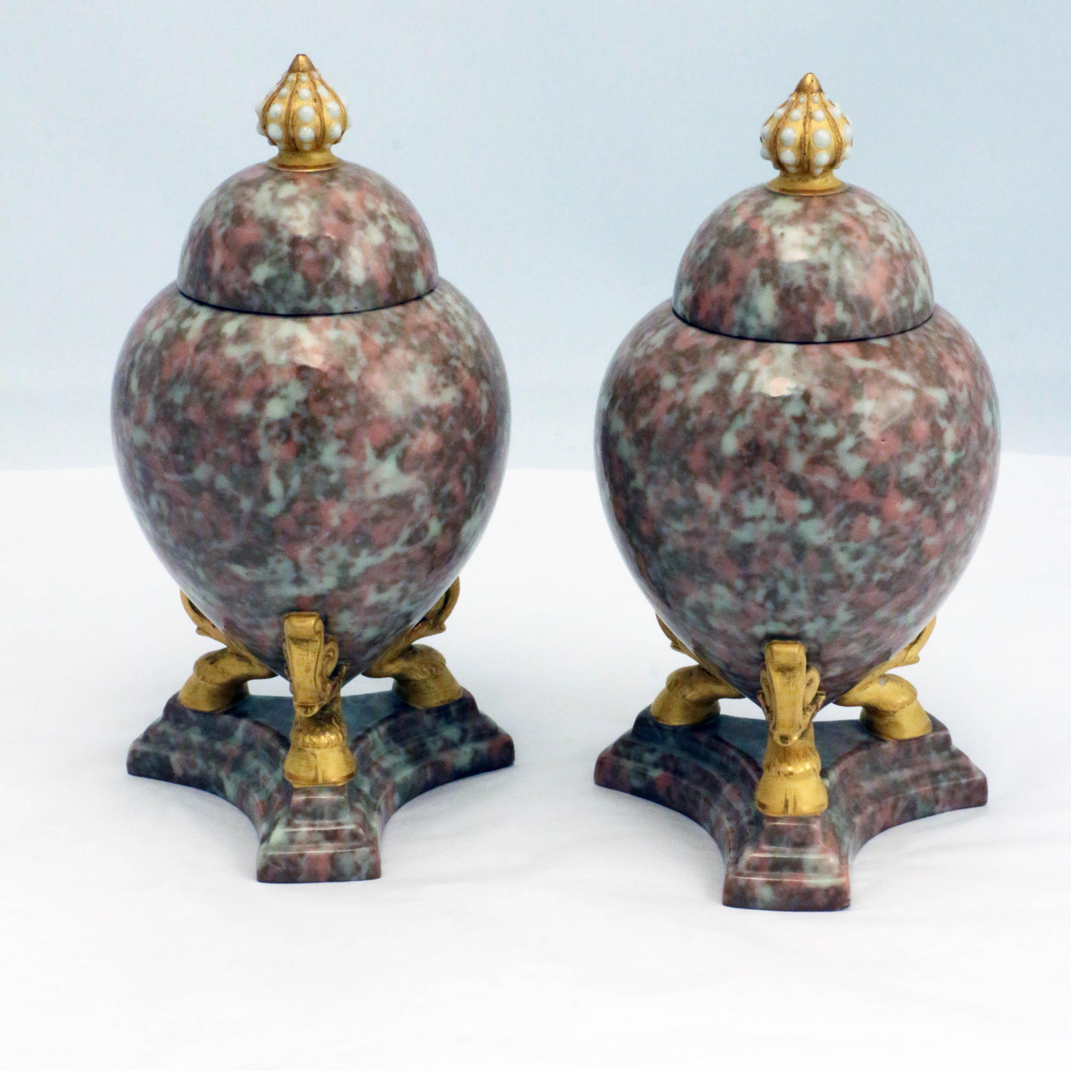 This pair of simulated marble covered urns are amphora-shaped, raised on gilt hoof feet on a stepped trefoil base, each domed cover has a lobed finial with 