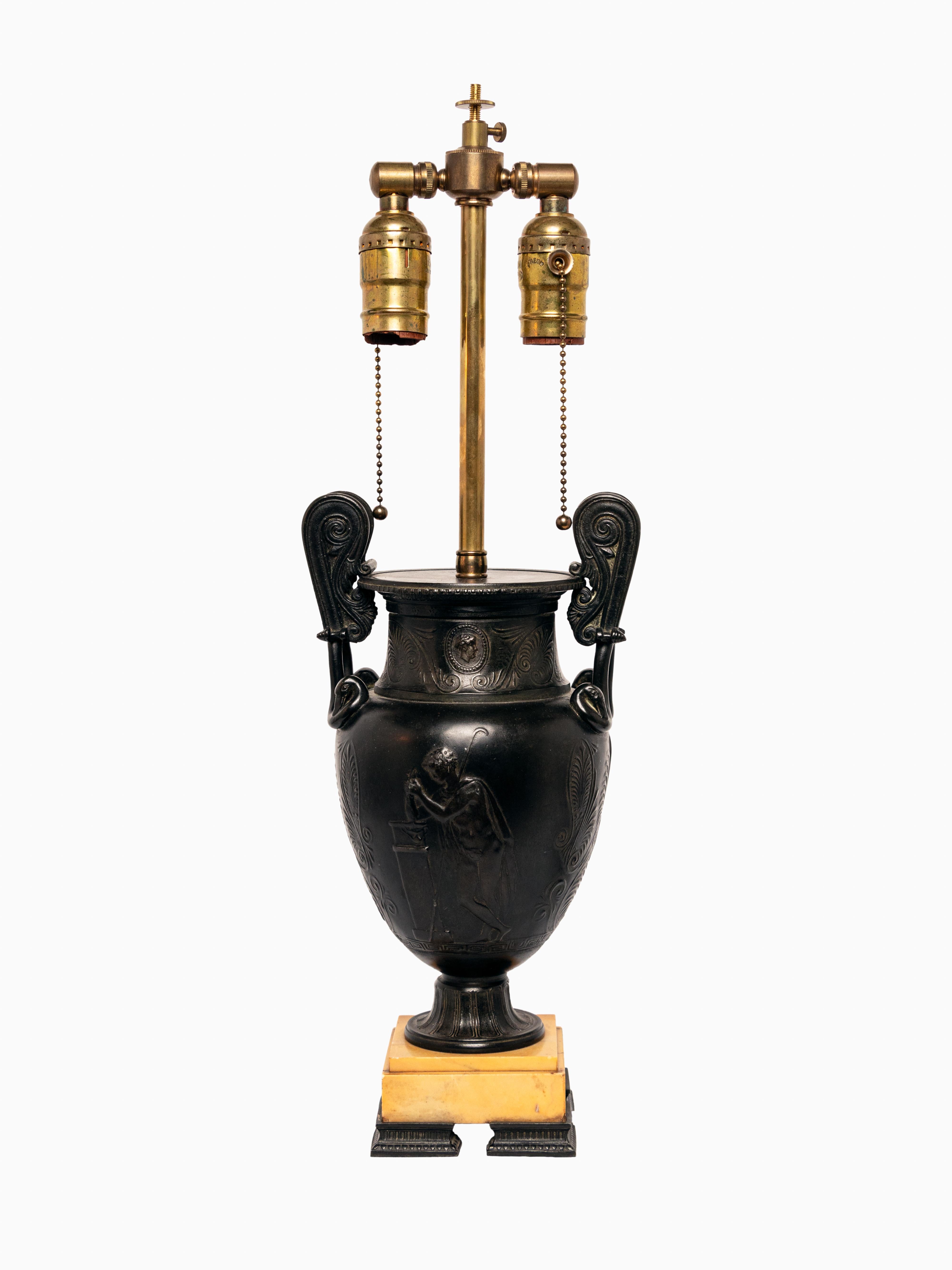 Paire d'urnes en bronze de la période du Grand Tour, montées comme lampes. L'urne en bronze est ornée d'images en bas-relief de figures classiques et de motifs feuillus. Les socles sont constitués de carrés de marbre ivoire et sont dotés de ferrures