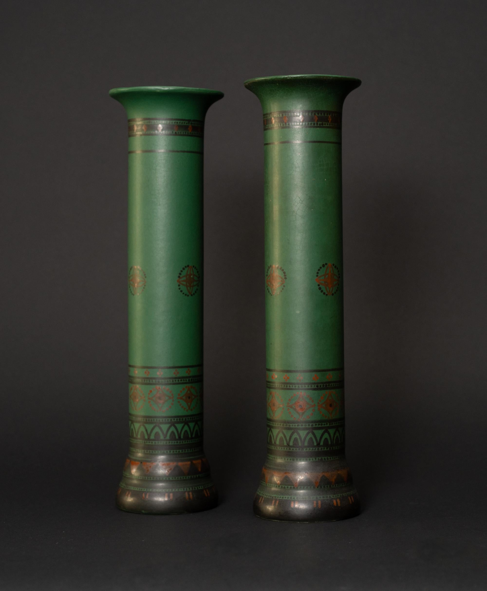 
These matching green pillar vases' bases are painted black and contain various geometric designs that work their way up the shaft of the vase, eventually becoming less concentrated. The designs utilize black, red, and gold motifs and generally use