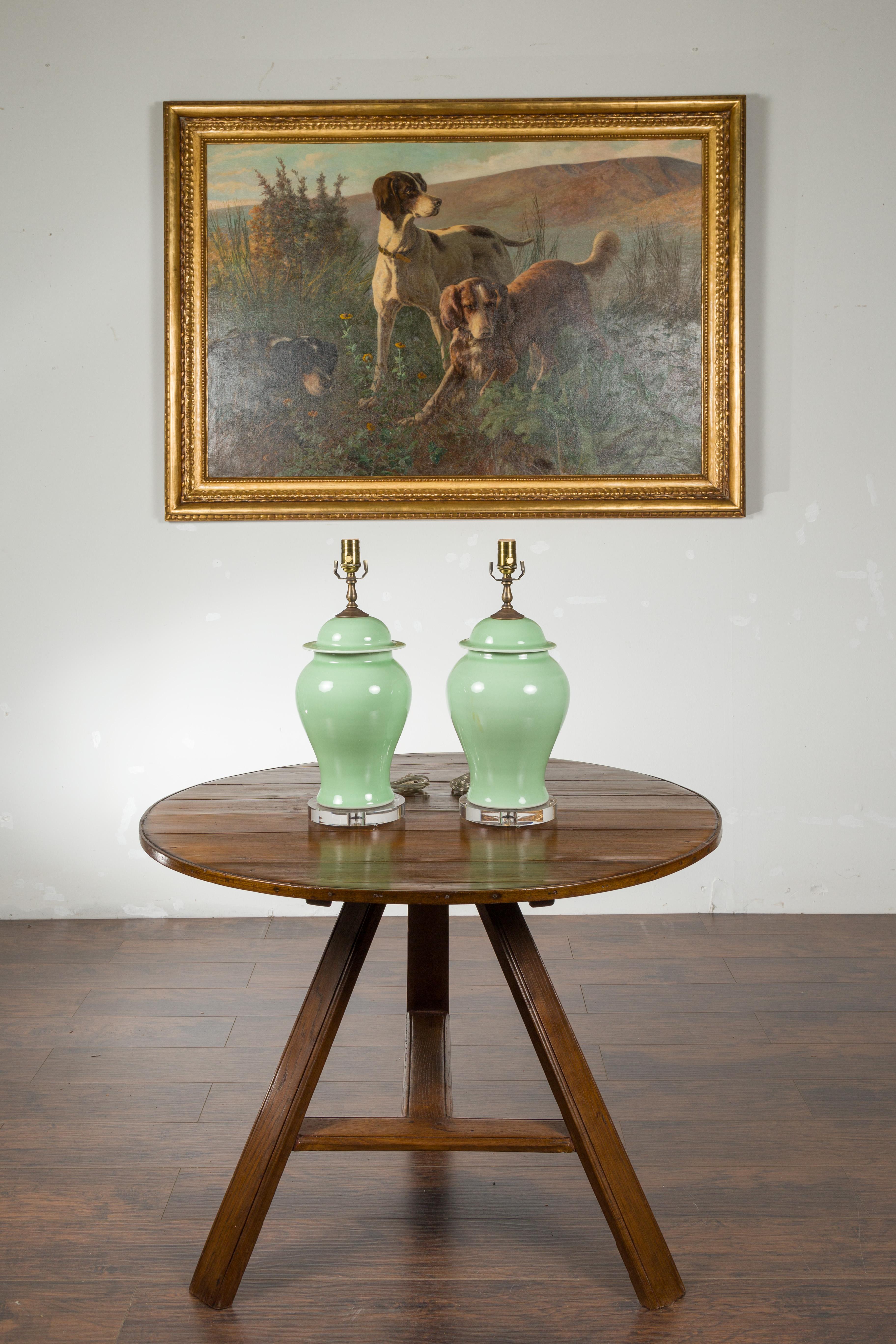 A pair of green porcelain lidded jar table lamps with lucite bases. Each of this pair of porcelain table lamps attracts our attention with its sleek lines and soft green color. Mounted on circular lucite bases and wired for the US, this pair of