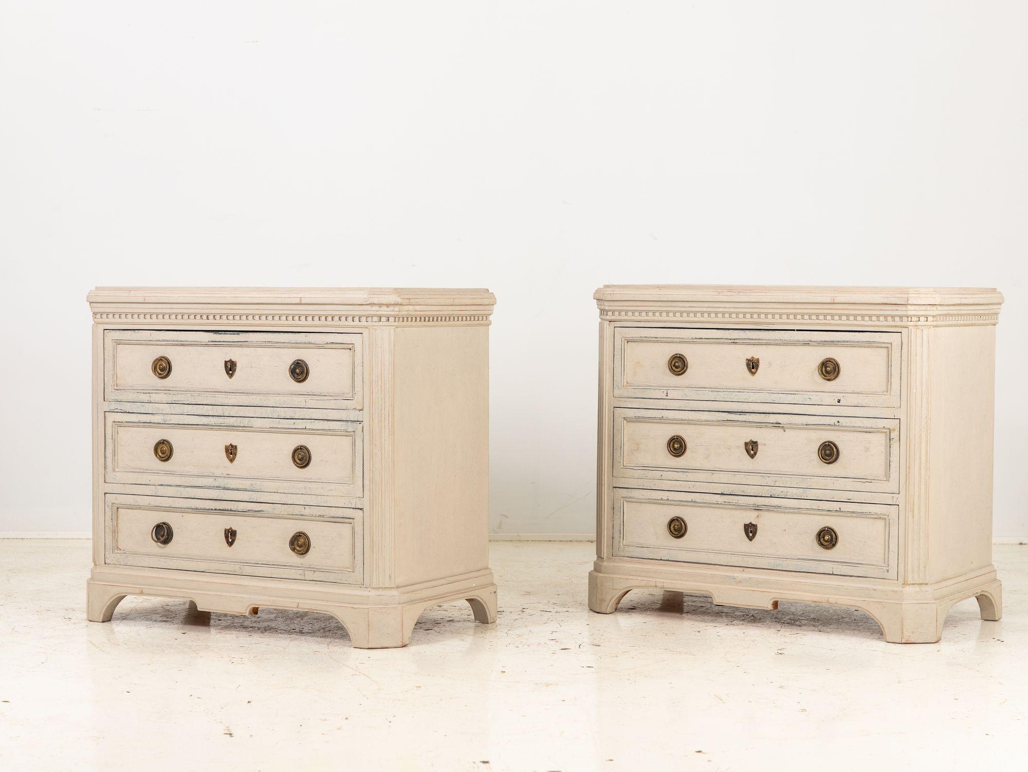 This pair of Gustavian style cream painted chests of drawers exudes timeless charm and sophistication. Each chest boasts three spacious drawers adorned with elegant brass ring pulls and escutcheons, adding a touch of opulence to their classical
