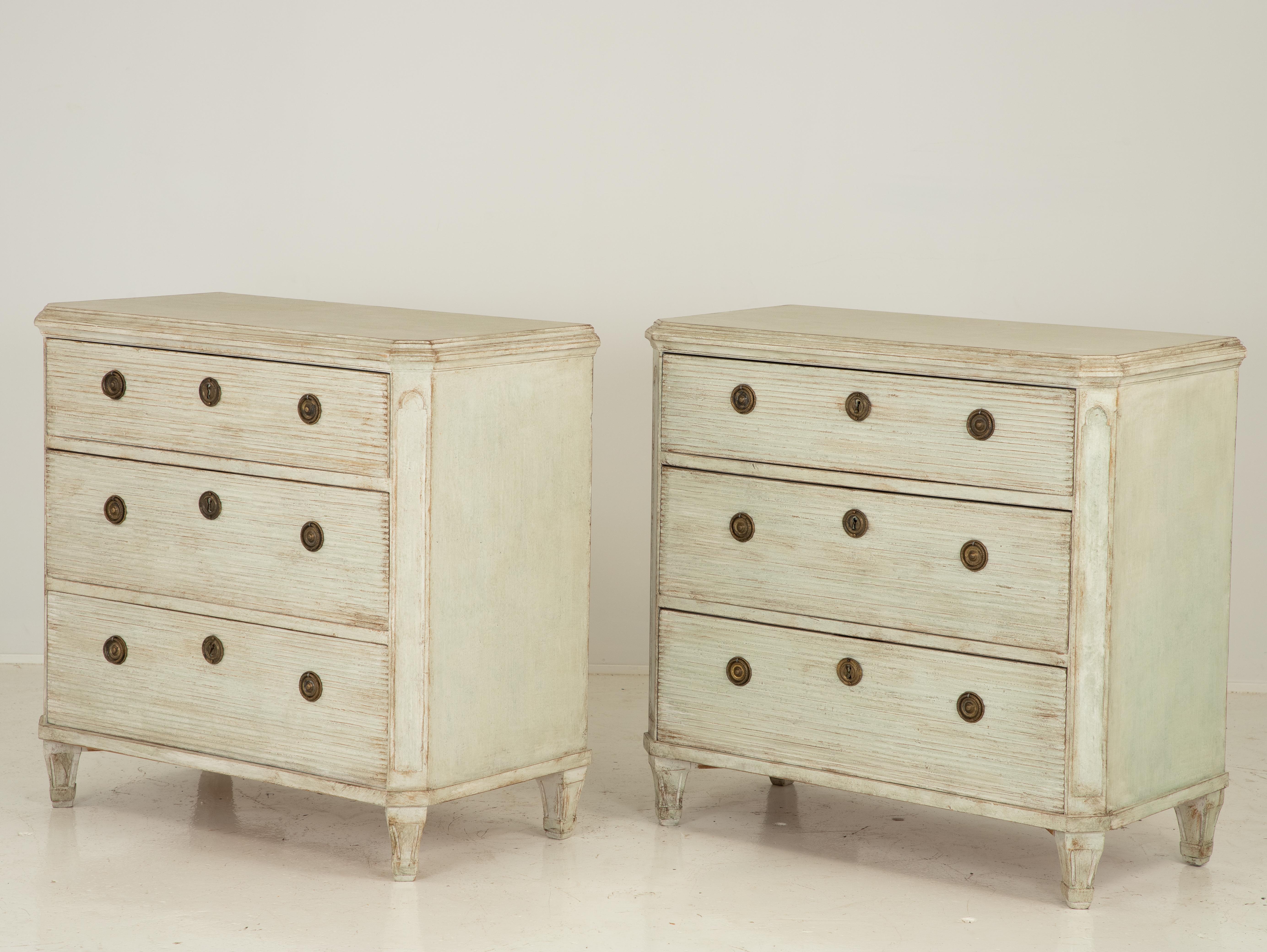 A late 19th century pair of painted Swedish Gustavian style commodes. Each commode has three drawers with brass ring pulls with beautifully carved reeded drawer fronts and two working keys. Canted corner on tapered feet. Dovetail joinery and later