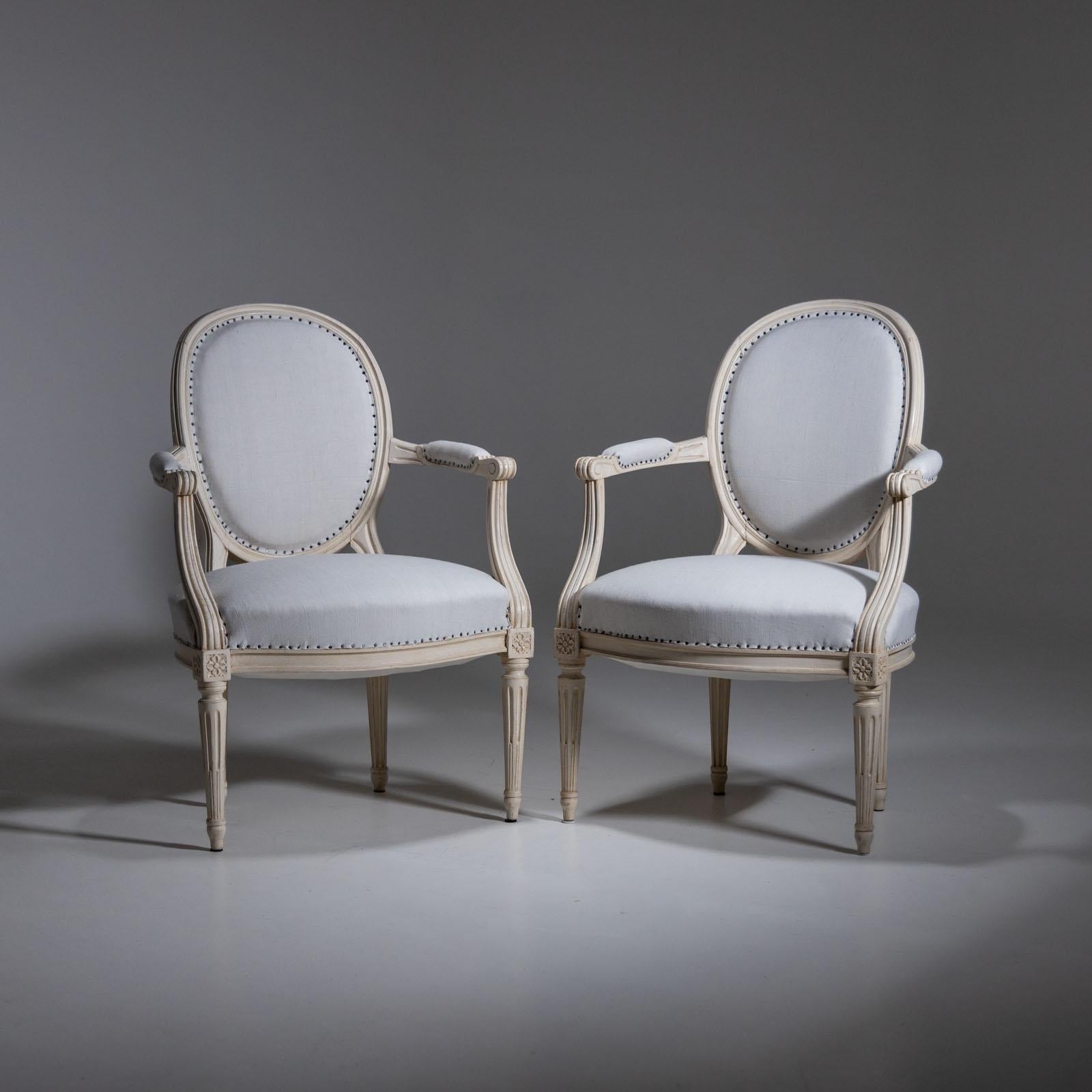 Pair of chairs with medallion-shaped upholstered backrests and upholstered armrests and seats. The beige paintwork is new and has a light antique patina. The chairs have been covered with an antique white linen fabric with studded decoration.
