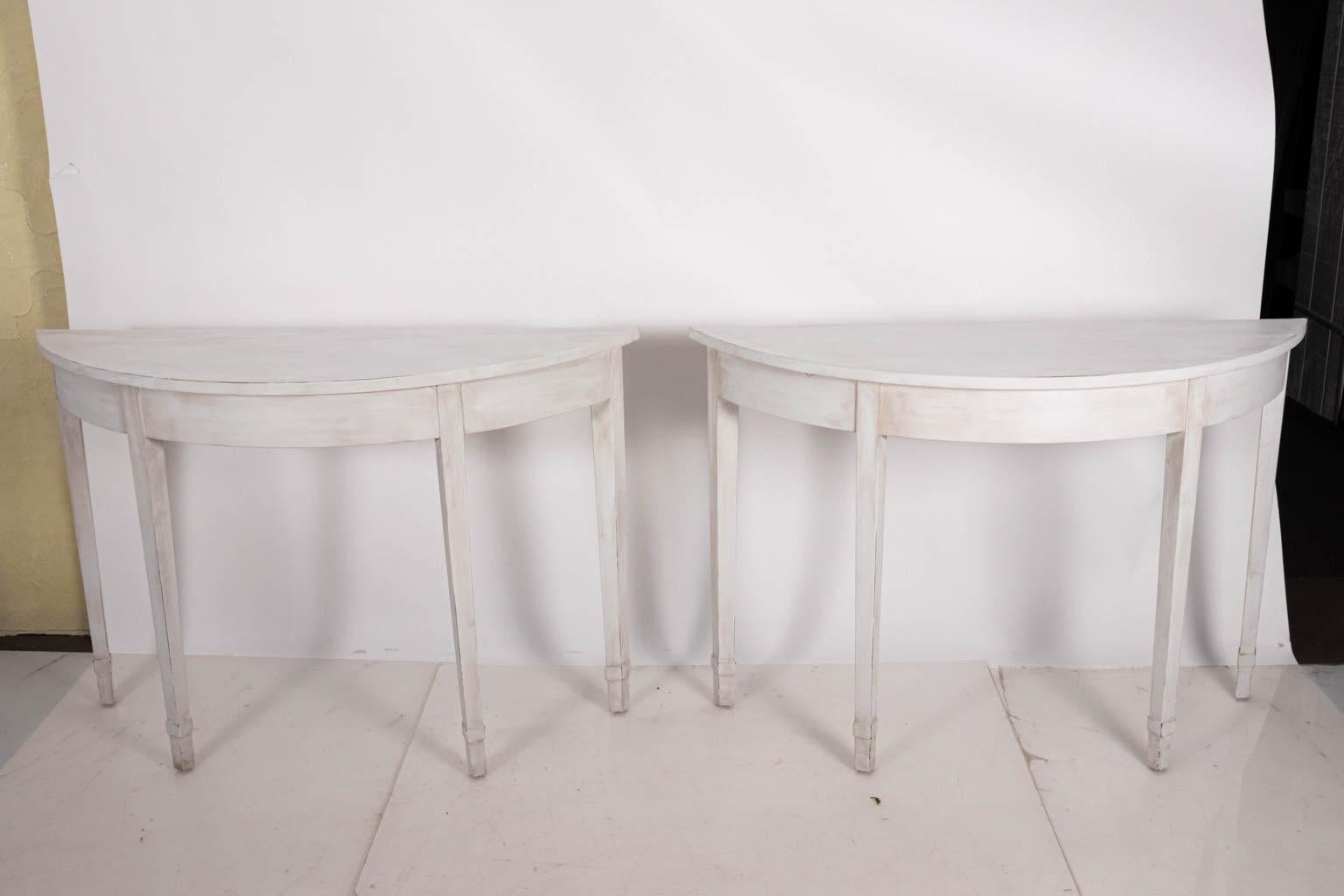 Pair of white painted demilune console tables in the Gustavian style, circa 1940s. The piece also features plain tapered legs and comes in an antique patina finish. Please note of wear consistent with age to the wood including: minor chips and dents