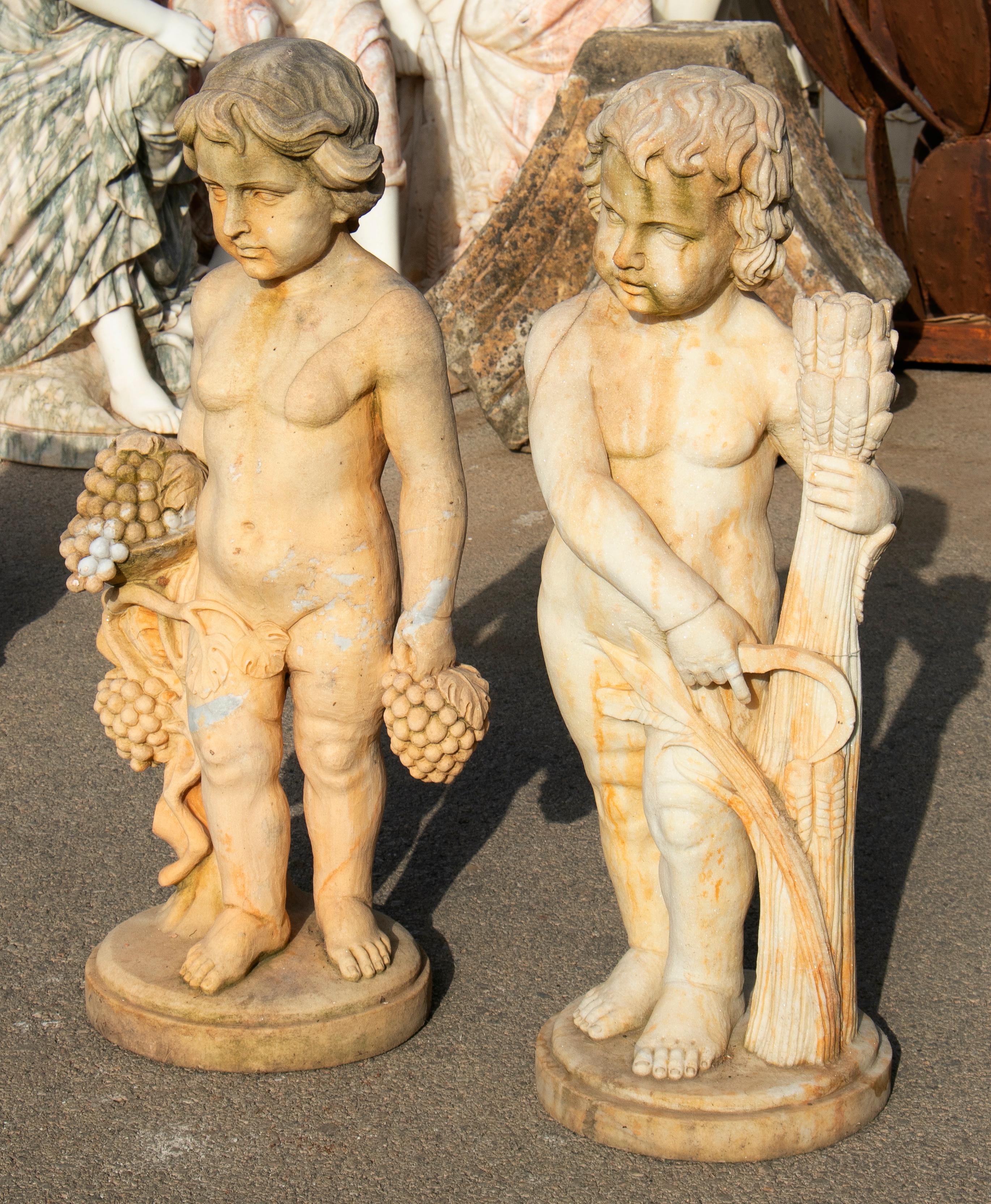 Pair of hand carved boys marble sculptures, one holding fruits and the other wheat, that have been aged to look antique.