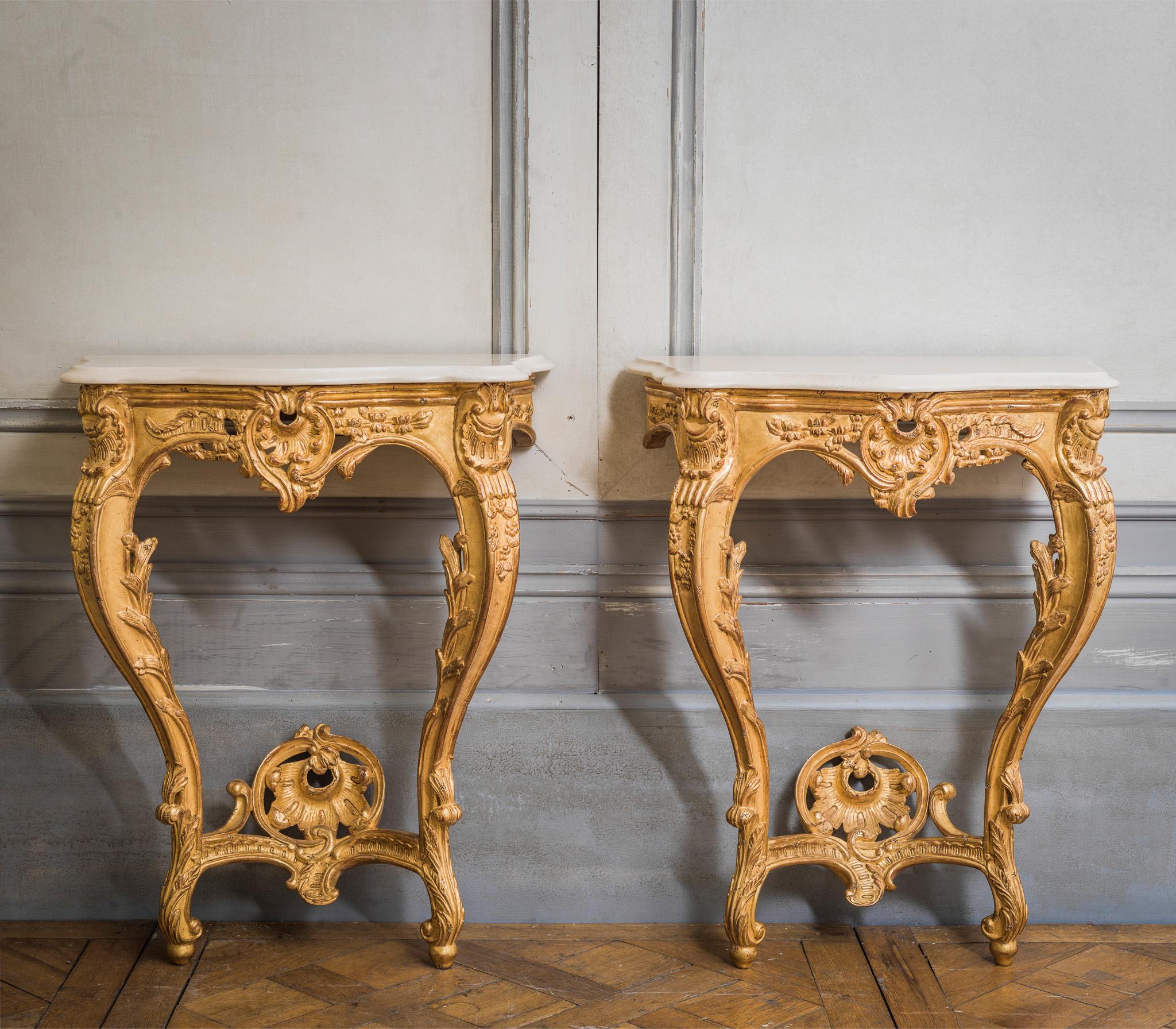 Louis XV style consoles hand-carved by master craftsmen and finished in a hand gilded patina, slightly aged and distressed. Including a bevel cut crema marfil marble which has been honed and lightly polished.