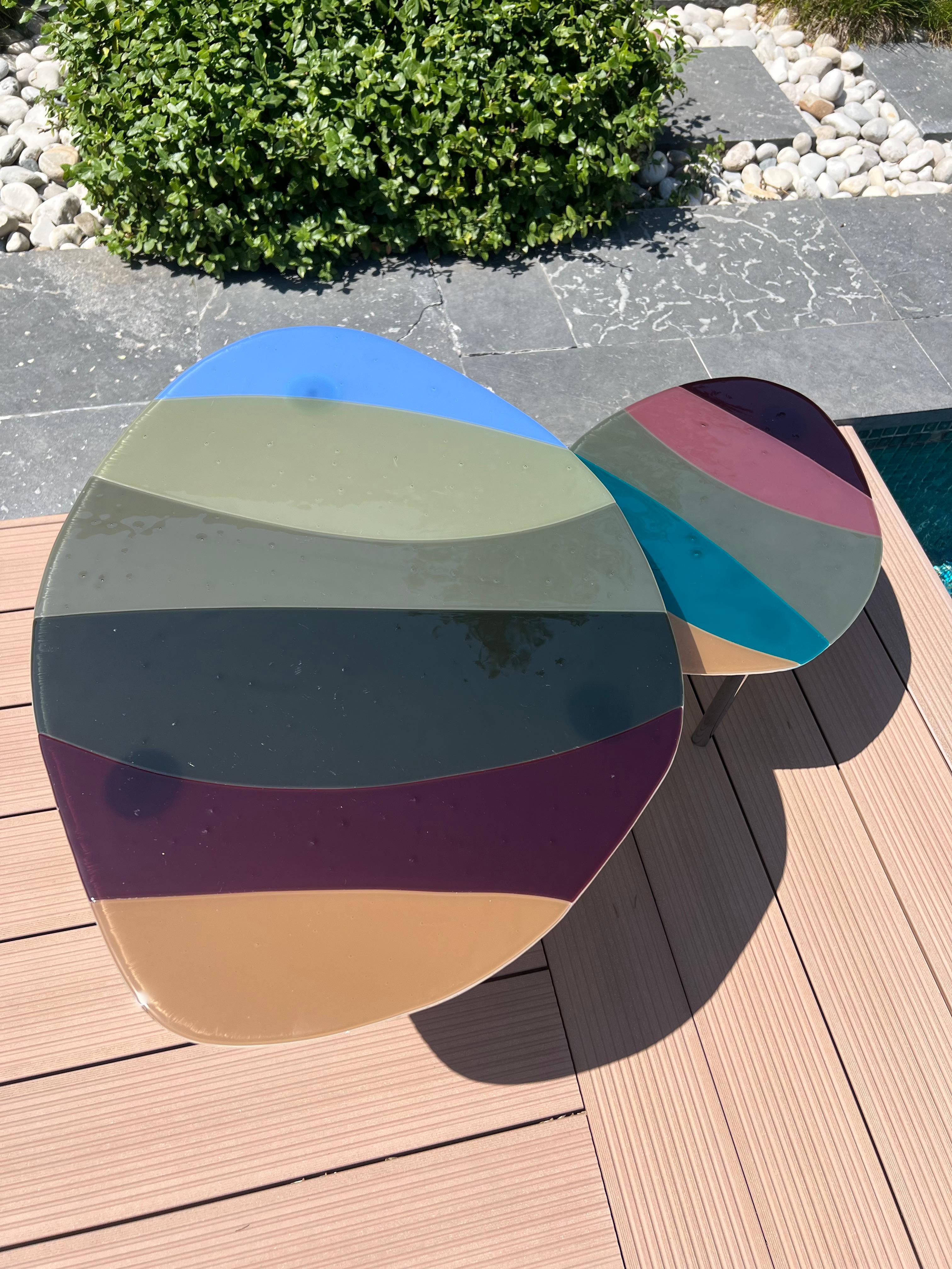 The two complementing nesting tables have fused glass surfaces and sleek metal legs.  Earth tones with splashes of blue and tourmaline will easily blend in. The pieces can be used indoors or outdoors.
The large table is 73 cm x 56 cm and 35 cm