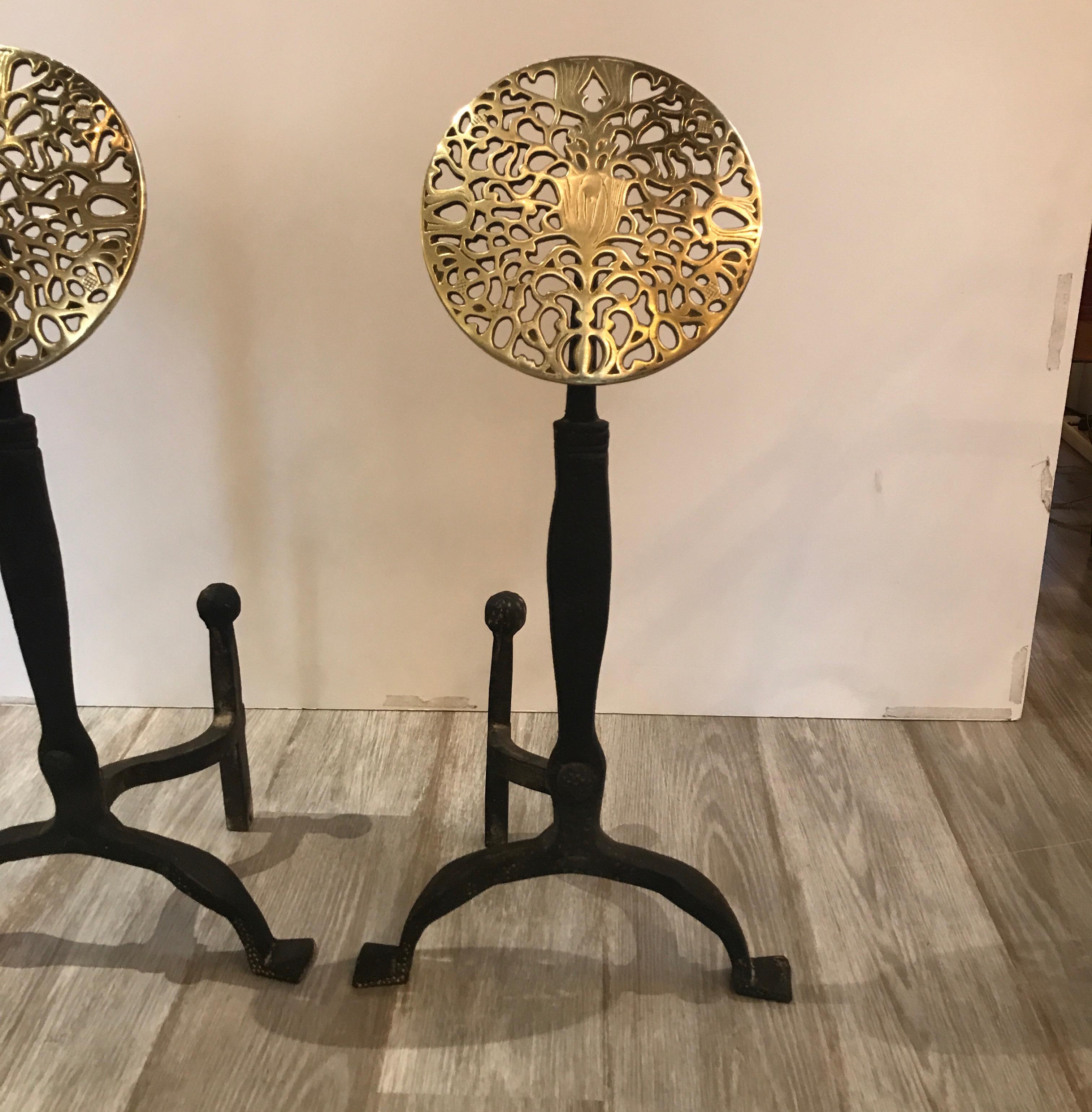 A pair of hand forged iron and polished brass Arts & Crafts andirons. A beautiful large pierced brass blossom medallion is the focal point. These are a short leg andiron that does not go back into the fireplace but are a Chenet that sits in the
