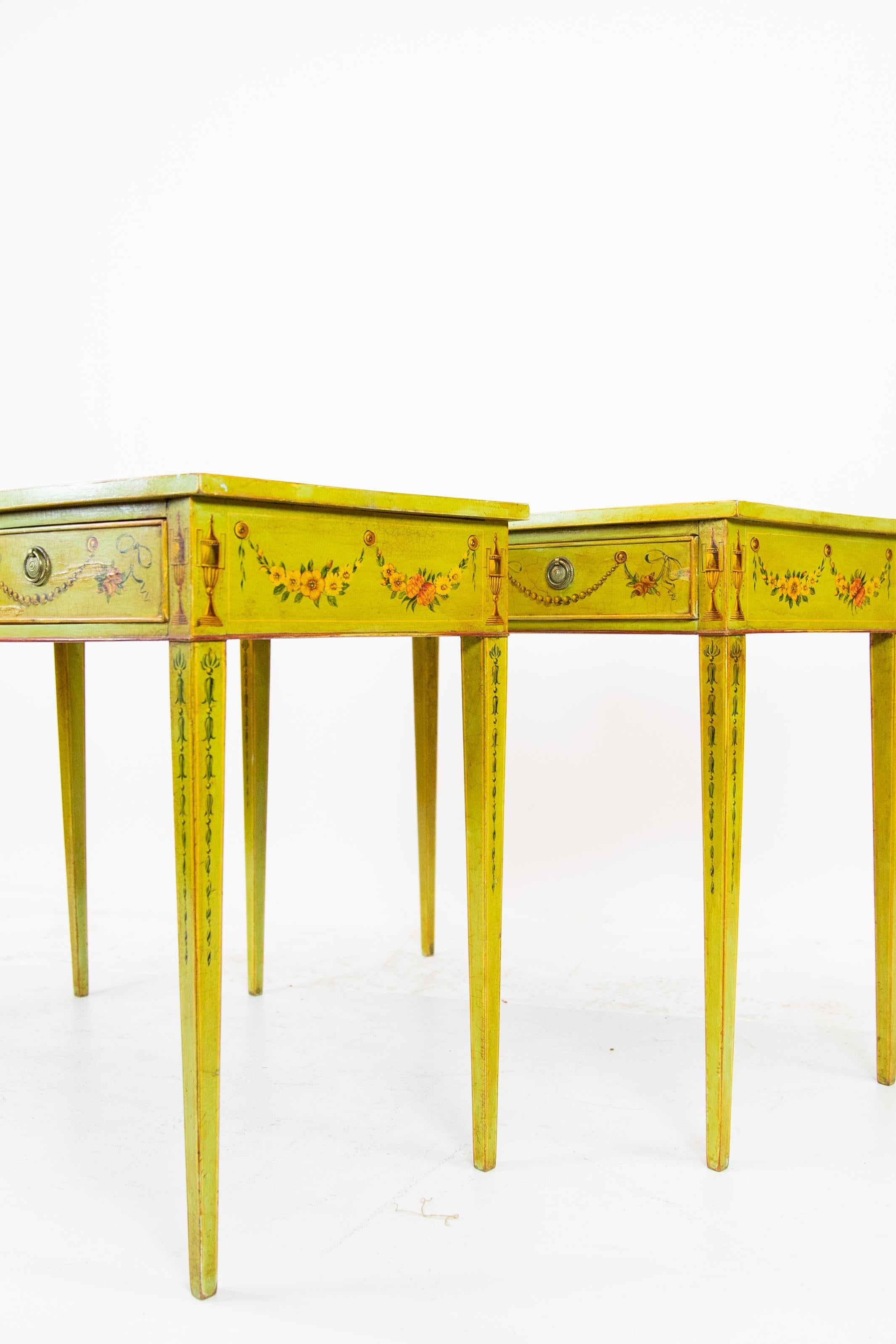 Coppia di tavolini dipinti a mano in bianco epple, con disegno floreale e di foglie. I dipinti raffigurano cestini floreali in un grande centrotavola, fasce trasversali con volute e strascichi di foglie. Urne classiche e campanule sono dipinte sulle