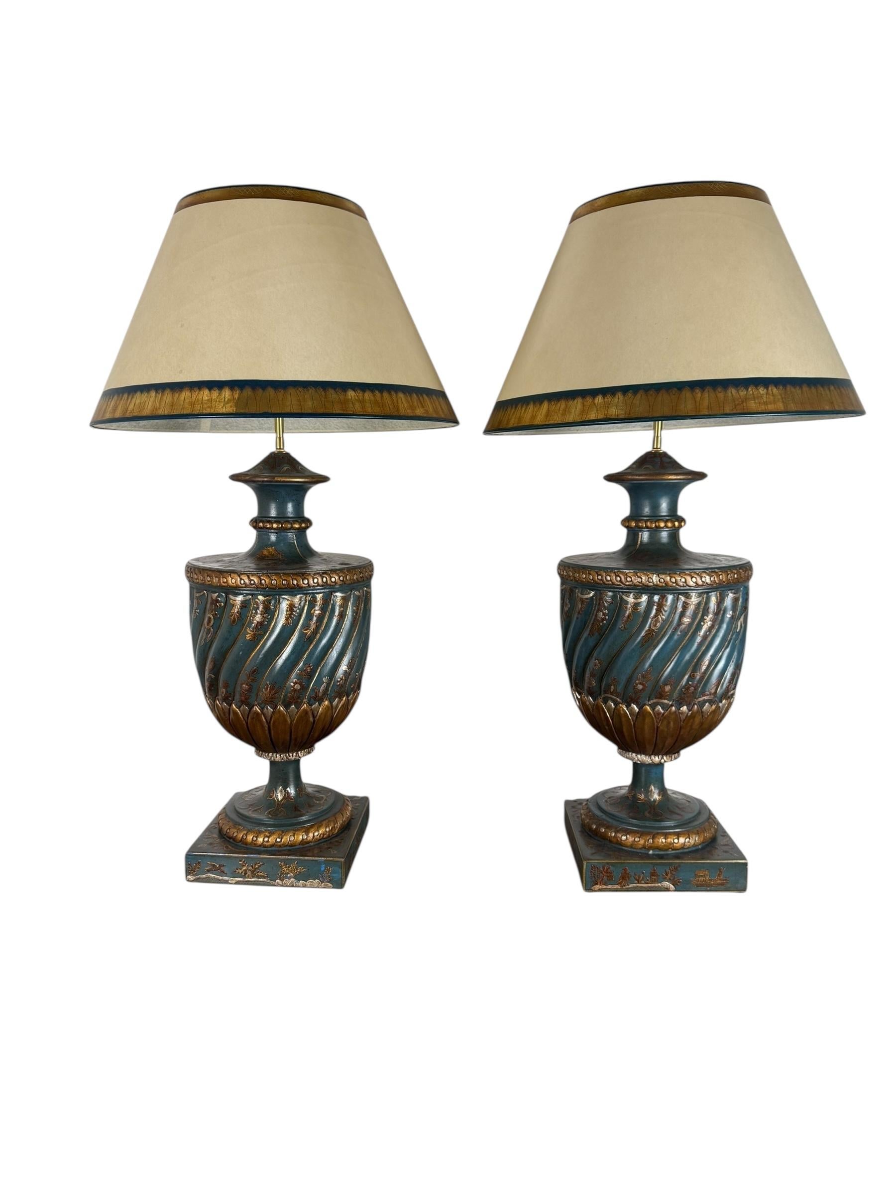 A striking pair of hand-painted urn-form table lamps in the chinoiserie taste, mounted on square plinth bases. The bodies are richly decorated with swirling teal ground and trailing gilt foliate motifs, accented with lotus-leaf borders and beadwork