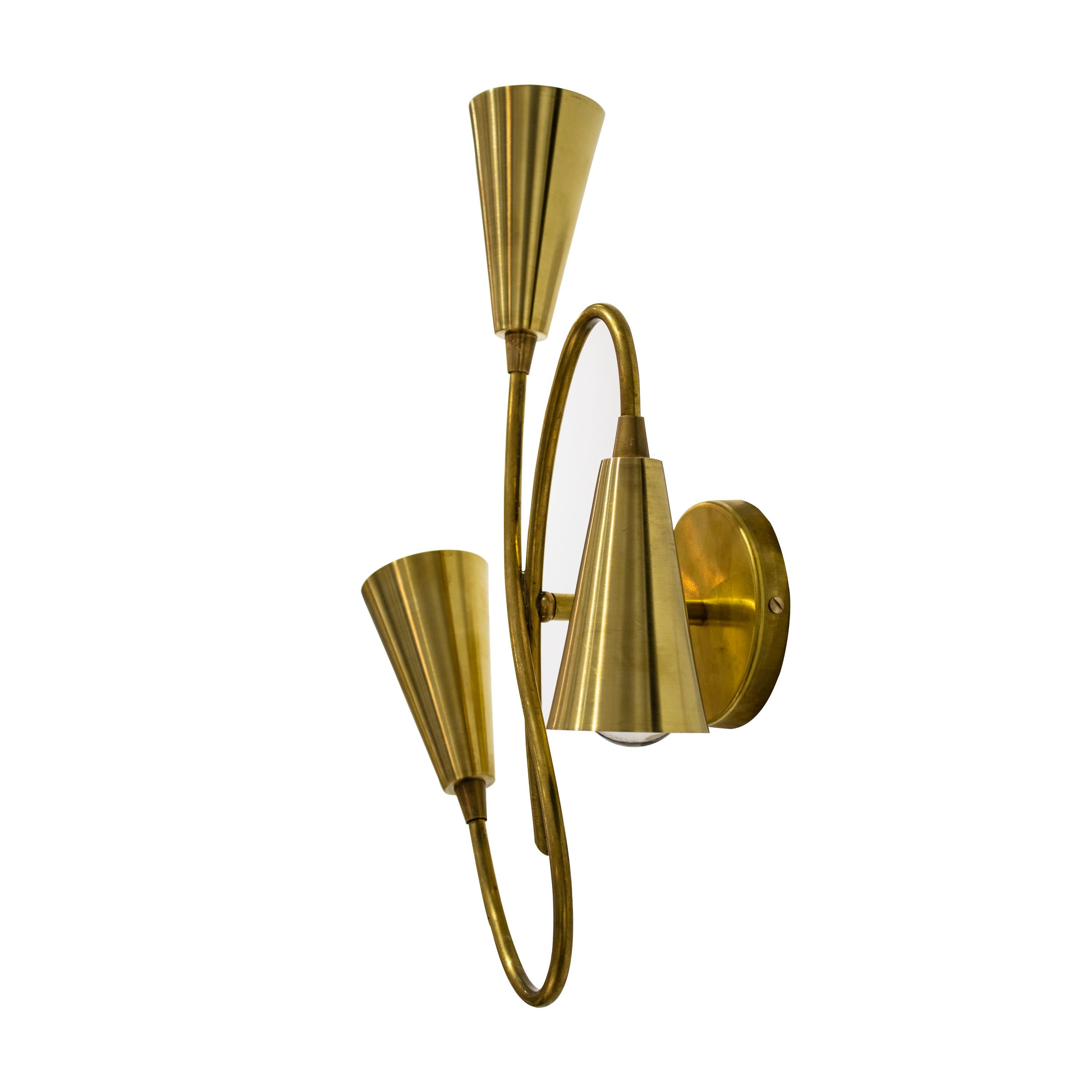 Pair of handcrafted Italian brass wall sconces. The lamps are made of curved brass tube structures with three light sources enclosed in cone lampshades in Stilnovo style. The lamp is attached to the wall through a round brass base. 



