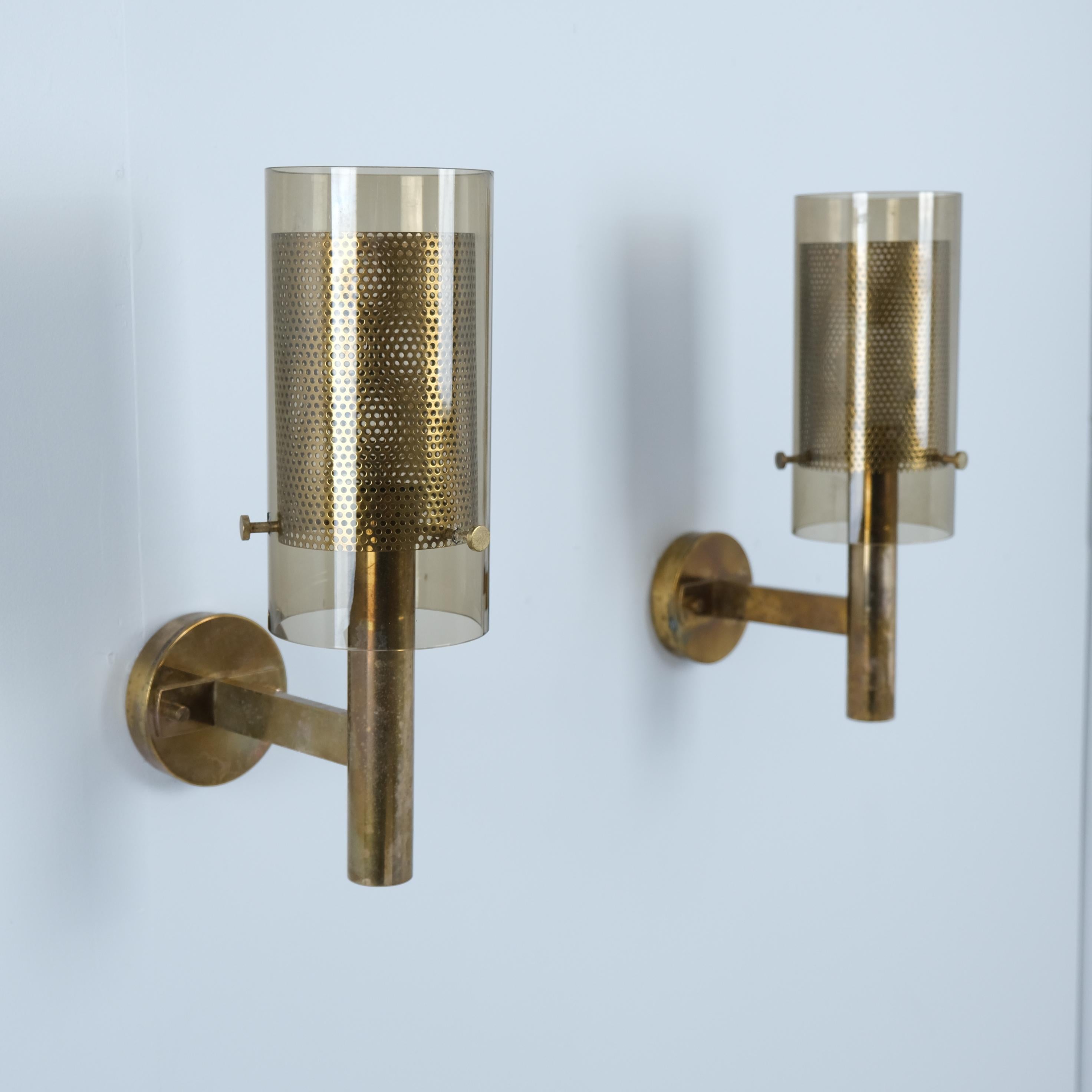 Una coppia di lampade da parete di Hans-Agne Jakobsson, modello V.I.I., che combinano il vetro fumé con un paralume interno in ottone perforato che diffonde la luce in un bagliore caldo e strutturato. Questa struttura a doppio strato è una delle