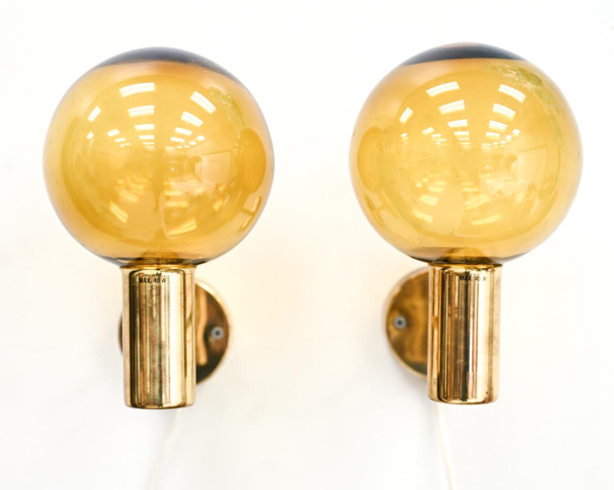 A pair of mid-century Scandinavian brass & glass electrified wall sconces designed by Hans-Agne Jakobsson for Markaryd (Sweden, 1960s). Polished brass with hand-blown amber glass spherical globe shades. Model V-149. With 