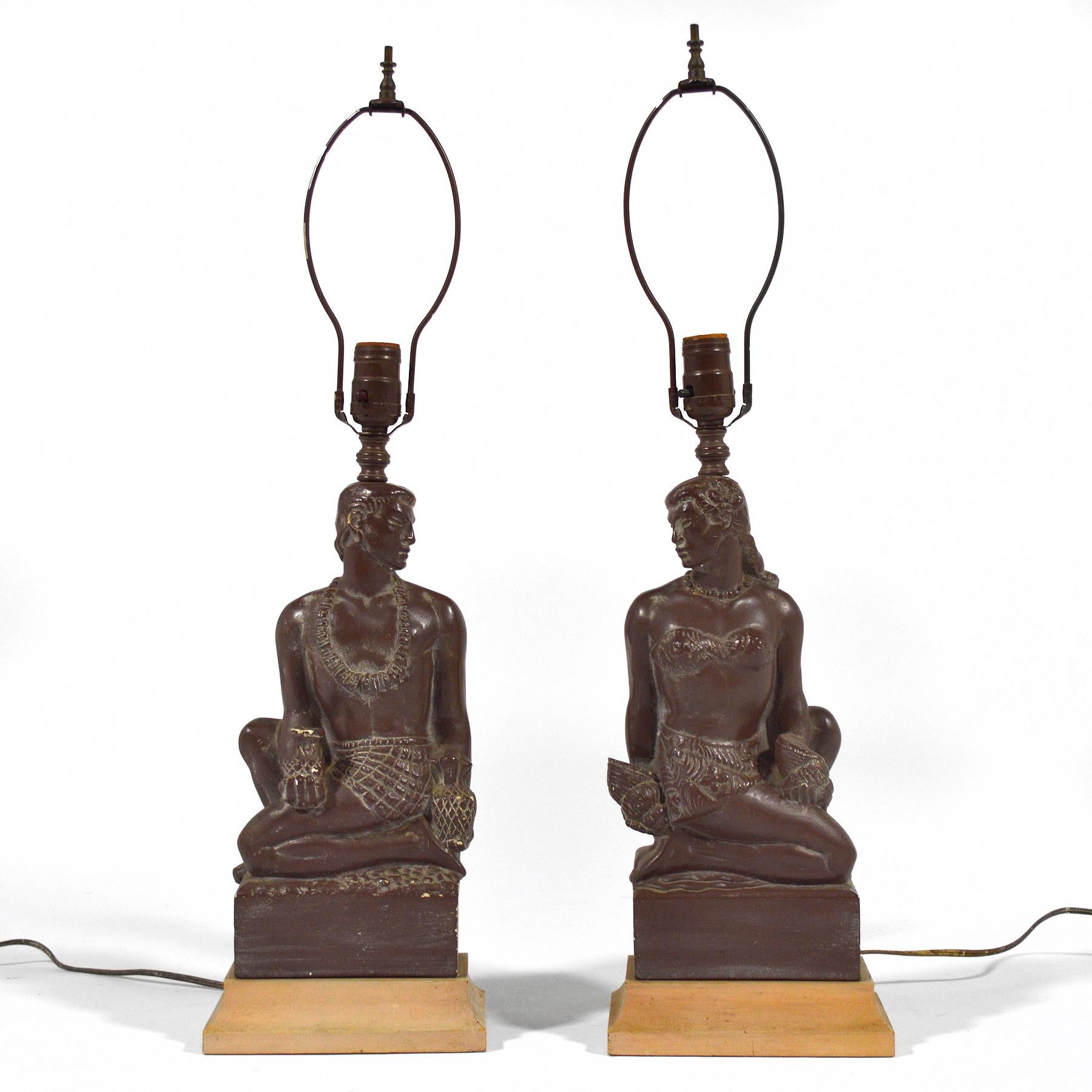 This fantastic pair of deco-inspired figural lamps feature a Hawaiian man and woman, are in good original condition, and have terrific style. The pair both wear leis and sarongs while the man holds a pair of pineapples and the woman has two shells.
