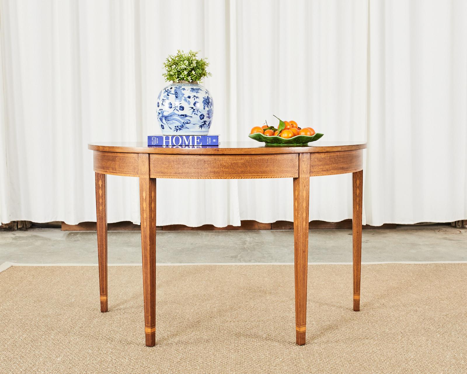 Distinctive pair of mahogany demi-lune tables or console tables crafted in the English Hepplewhite taste. The consoles feature delicate satinwood inlay with thread accents and bell flowers on the frieze and legs. Supported by elegant, tapered legs